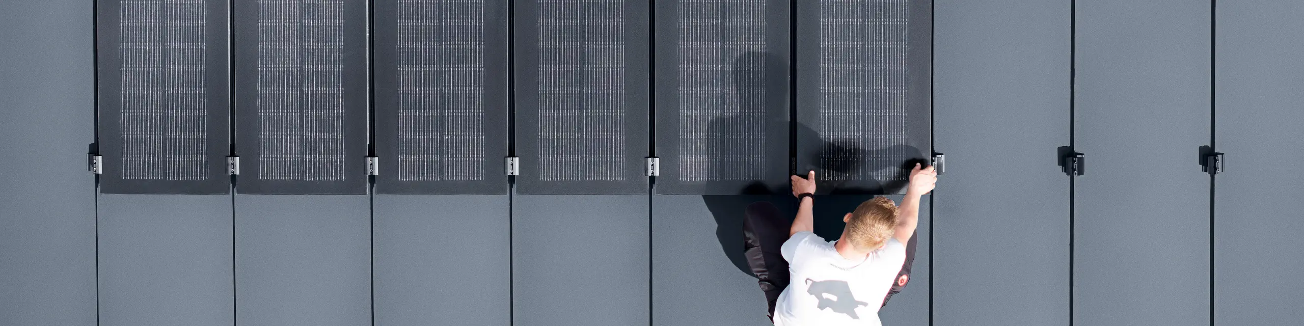 Zwei Personen installieren PREFALZ-Solarmodule auf einer großen grauen Dachfläche, die mit doppelflanschigen Paneelen bedeckt ist.
