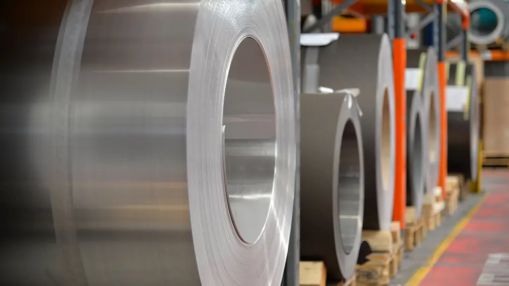 Aluminiumsruller (spoler) oplagret på træpaller, i forgrunden står en naturblank aluminiumsrulle, og bagved dem ses flere bånd og plader af aluminium.