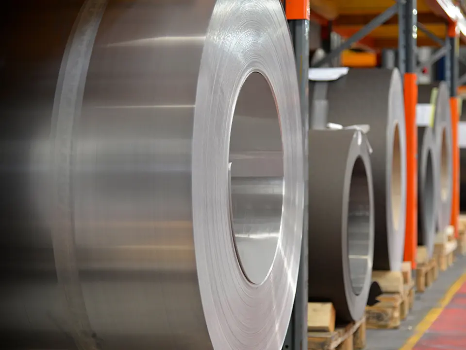 Aluminiumsruller (spoler) oplagret på træpaller, i forgrunden står en naturblank aluminiumsrulle, og bagved dem ses flere bånd og plader af aluminium.