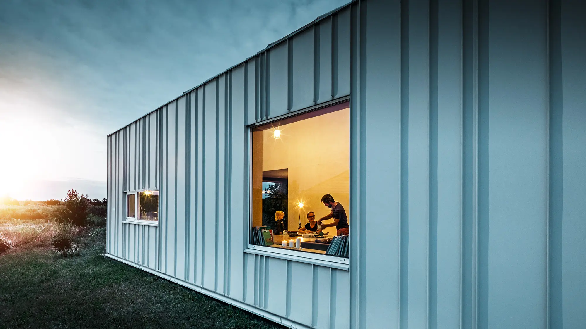 Vinkeltstående falsfacade med PREFALZ i forskellige panelbredder i P.10-prefahvid fotograferet i skumringen. I spisestuen kan man se en familie spise middag. 