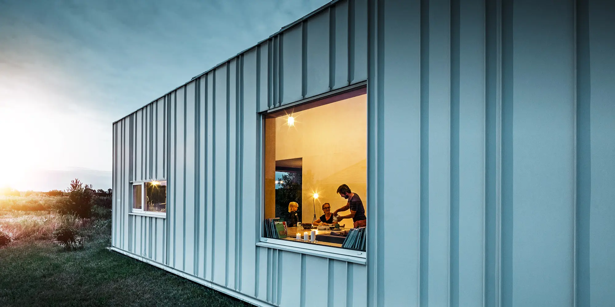 Vinkeltstående falsfacade med PREFALZ i forskellige panelbredder i P.10-prefahvid fotograferet i skumringen. I spisestuen kan man se en familie spise middag. 