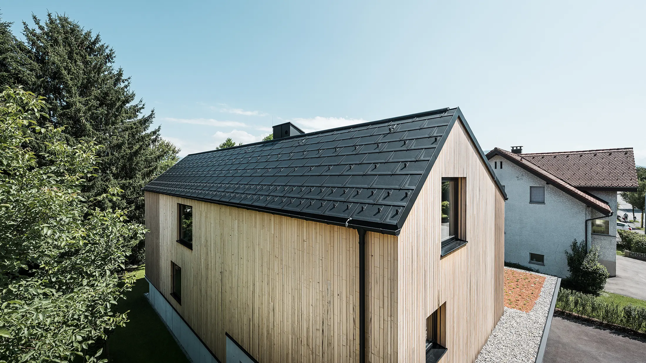 Ein modernes Einfamilienhaus in Lustenau besticht durch die Kombination einer freundlichen Holzfassade mit robusten PREFA Aluminiumprodukten. Das Satteldach wird von der PREFA Dachplatte R.16 mit Schneestoppern sowie der passenden Dachentwässerung in Form von Hängerinne und Ablaufrohr geschmückt. Alle Produkte sind einheitlich im Farbton Schwarz P.10 gehalten und harmonieren perfekt mit der vertikalen Holzfassade.