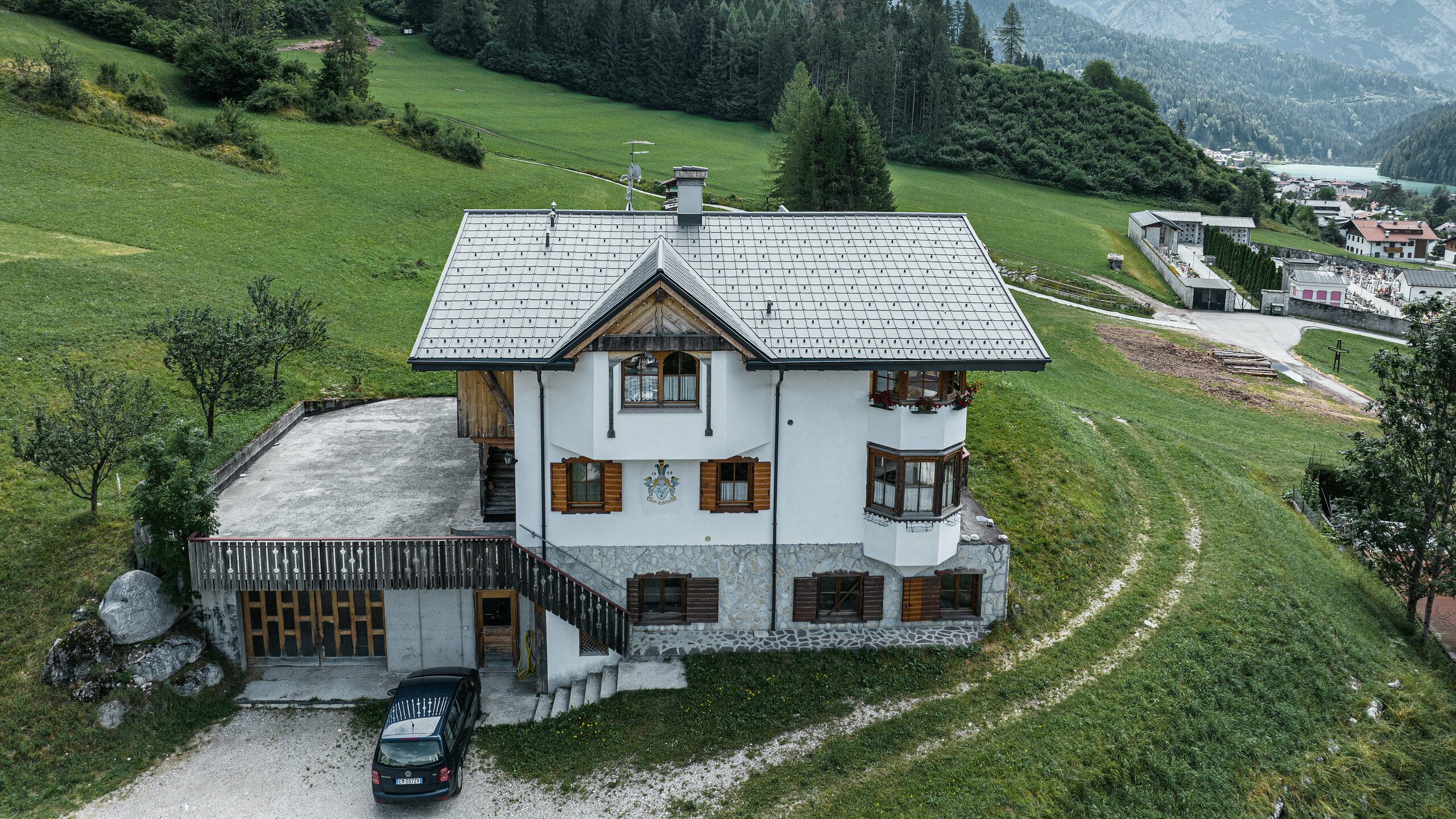 Fugleperspektiv af indgangen til den traditionelle bjerghytte i Auronzo di Cadore efter tagrenoveringen med PREFA aluminiumsfliser i P.10 lys grå. Bygningen med sin karakteristiske alpine charme og moderne bliktag falder harmonisk ind i det frodige grønne landskab og det imponerende bjergpanorama i baggrunden og viser en vellykket kombination af funktionalitet og regional arkitektur.
