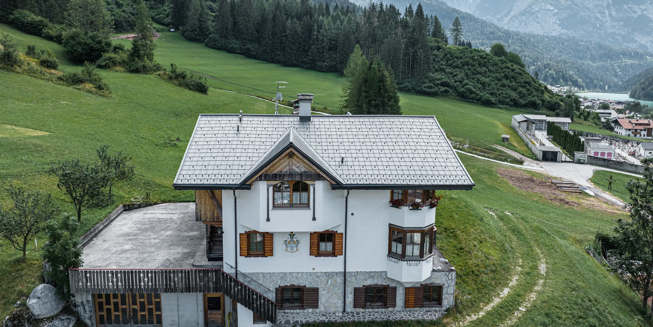 Fugleperspektiv af indgangen til den traditionelle bjerghytte i Auronzo di Cadore efter tagrenoveringen med PREFA aluminiumsfliser i P.10 lys grå. Bygningen med sin karakteristiske alpine charme og moderne bliktag falder harmonisk ind i det frodige grønne landskab og det imponerende bjergpanorama i baggrunden og viser en vellykket kombination af funktionalitet og regional arkitektur.