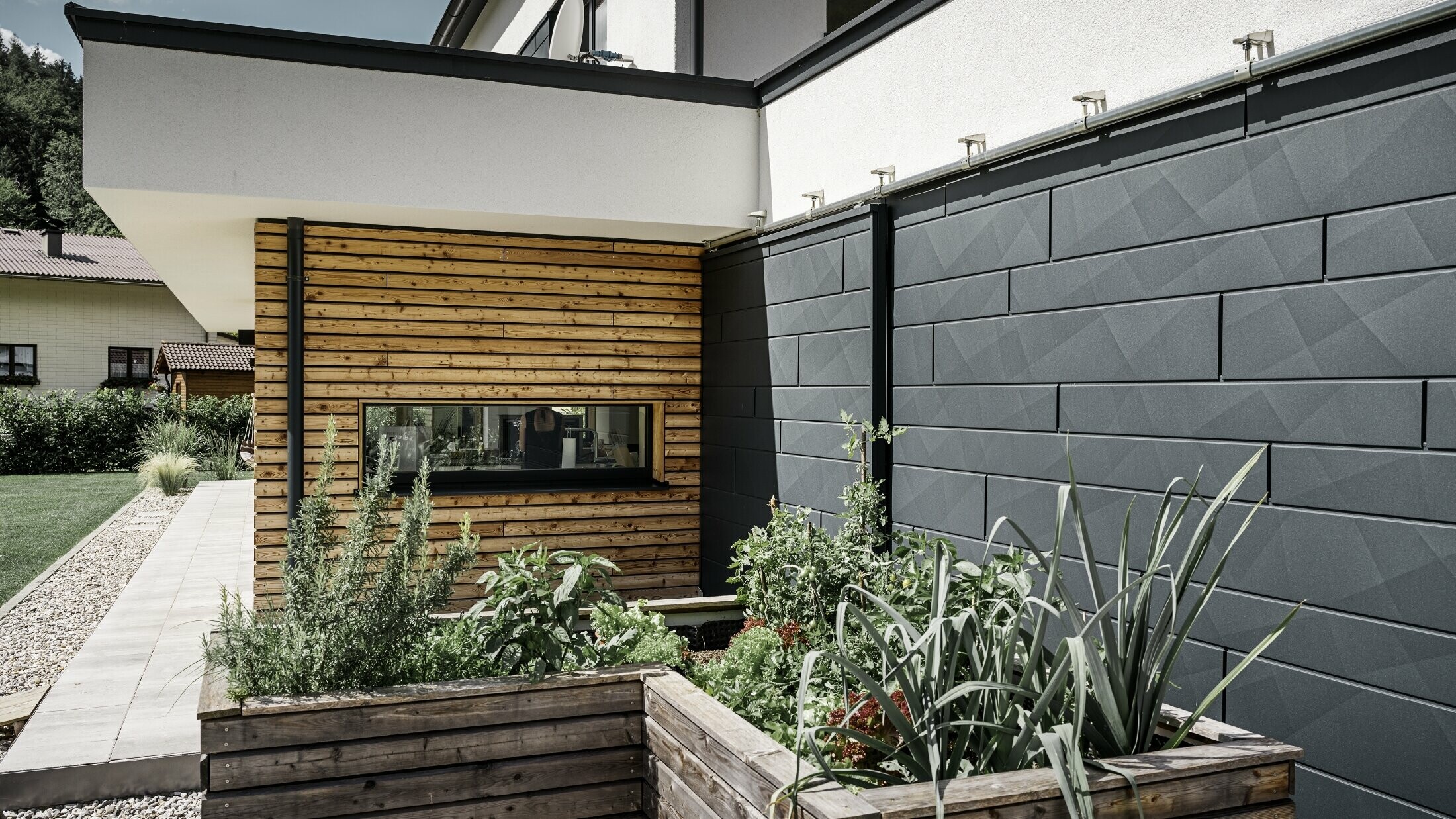 Dette billede viser et nærbillede af enfamiliehuset, hvor fokus er på facadebeklædningen med PREFA Siding.X i P.10 antracit. De mørke facadepaneler med det markante krøllede udseende skaber en flot visuel kontrast til de livlige, træbeklædte områder og de omgivende grønne områder. Foran den facade af aluminium er der placeret højbede, der er beplantet med forskellige planter og buske, hvilket understreger husets naturlige og bæredygtige udstråling. Kombinationen af moderne materialer og integrationen i haveområdet formidler et billede af moderne arkitektur, der harmonisk indpasser sig i omgivelserne.