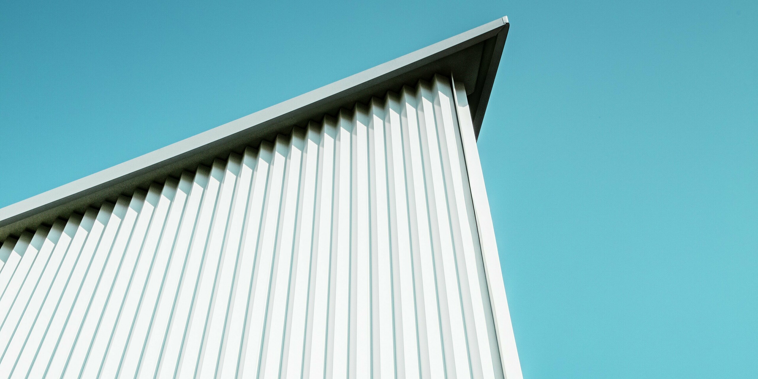 Dramatische Perspektive auf die markante PREFA Fassade mit Zackenprofil in P.10 Reinweiß vor klarem Himmel. Die präzise Linienführung der Aluminiumverkleidung unterstreicht die architektonische Schärfe. Das witterungsbeständige und langlebige Blech sorgt für höchste Stabilität bei minimalem Pflegeaufwand – perfekt für alle Gebäudetypen.