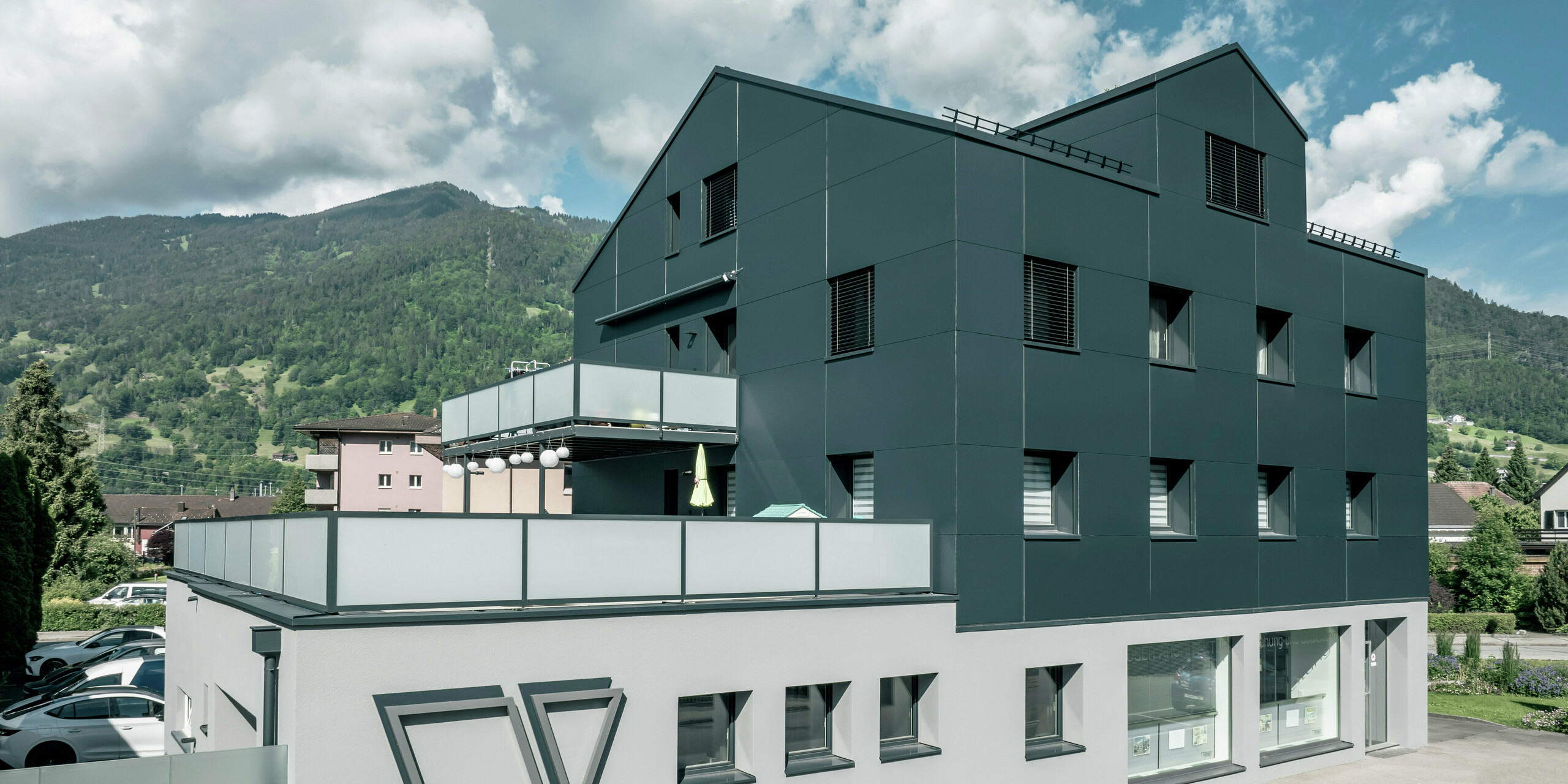 Das Architekturbüro Voser in Landquart, im Schweizer Graubünden, mit einer PREFA Aluminiumfassade in Anthrazit. Das Gebäude wurde saniert und mit PREFABOND Aluminium Verbundplatten ausgestattet. Die Gestaltung kombiniert klare Linien und hochwertige Materialien. Auffällig sind die großen Balkone mit Glasbrüstungen und das prägnante Logo des Architekturbüros Voser an der Gebäudeseite. Im Hintergrund ist eine grüne Berglandschaft zu erkennen.
