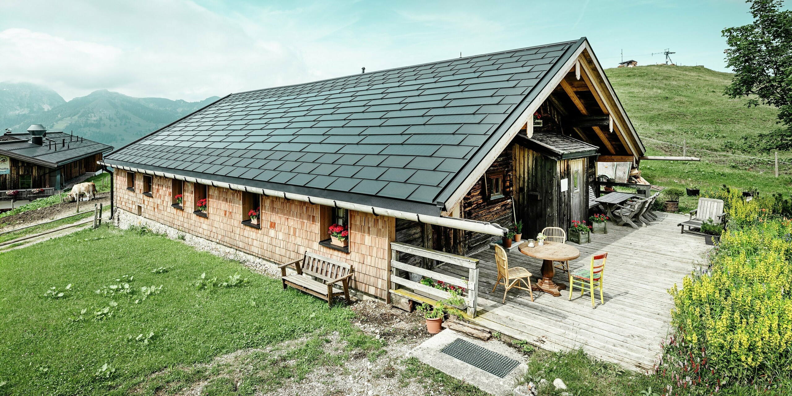 Vorderansicht der Birkehütte auf der Walleralm in Bayrischzell, eingebettet in die malerische Bergwelt. Das Dach wurde im Zuge einer Sanierung mit innovativen PREFA Solardachplatten ausgestattet. Das moderne Dachsystem kombiniert ein langlebiges Aluminiumdach mit einer integrierten Photovoltaikanlage. Ergänzt wird die Dachfläche durch robuste PREFA Dachplatten R.16 in der Farbe P.10 Schwarz. Die Kombination aus moderner Photovoltaik-Technologie und traditioneller Holzbauweise sorgt für nachhaltigen Wetterschutz und fügt sich harmonisch in die alpine Umgebung ein. Im Vordergrund des Bildes ist die einladende Terrasse mit rustikalen Holzmöbeln zu sehen. 