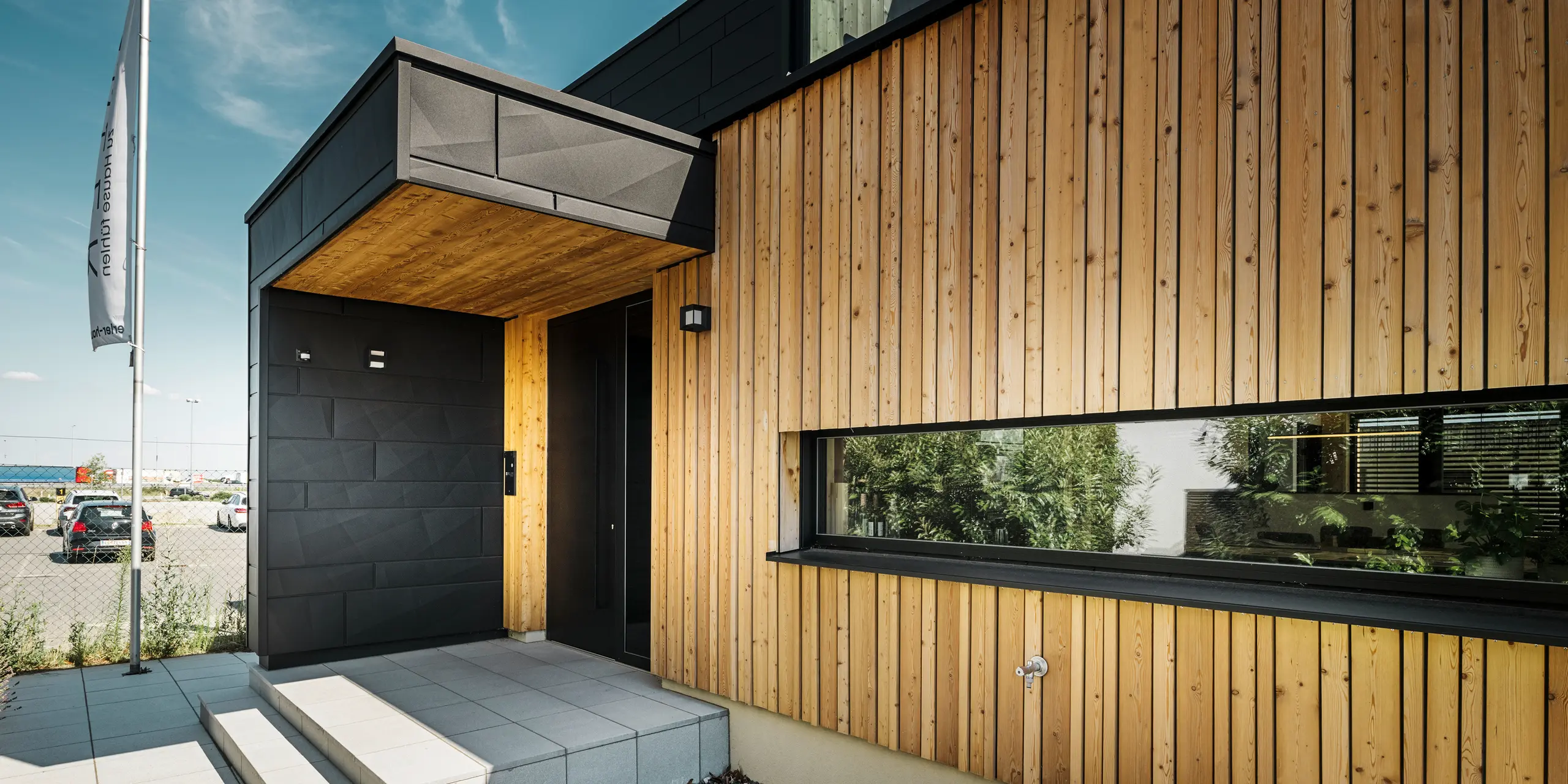 Sideindgang til Autark-udstillingshuset i Blaue Lagune præfabrikerede huscenter i Vösendorf med en moderne facade af naturligt træ og sorte PREFA Siding.X-paneler. Billedet viser den elegante kombination af træbeklædning og aluminium i højkvalitetsbelægningen P.10 Black.