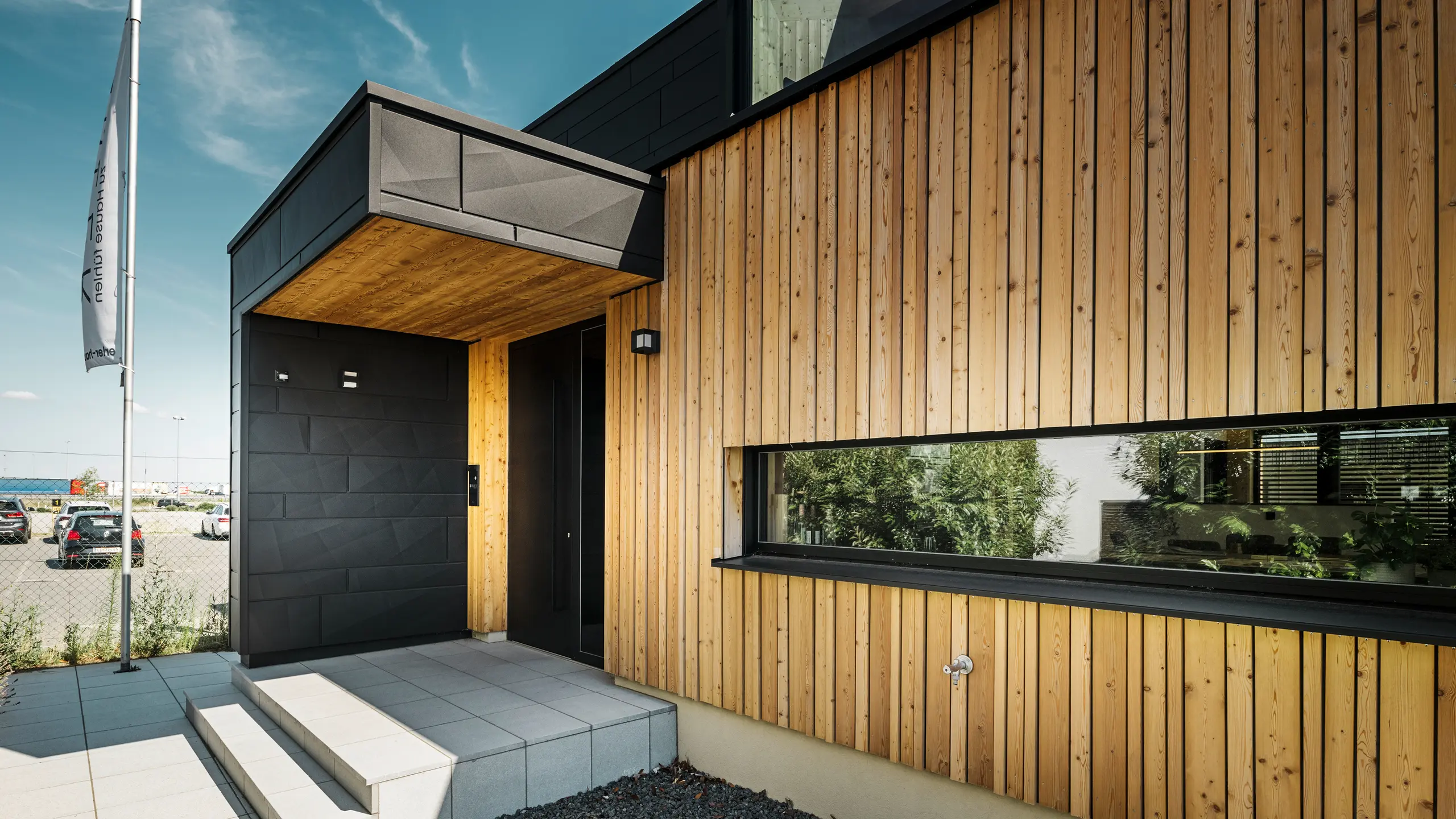 Sideindgang til Autark-udstillingshuset i Blaue Lagune præfabrikerede huscenter i Vösendorf med en moderne facade af naturligt træ og sorte PREFA Siding.X-paneler. Billedet viser den elegante kombination af træbeklædning og aluminium i højkvalitetsbelægningen P.10 Black.