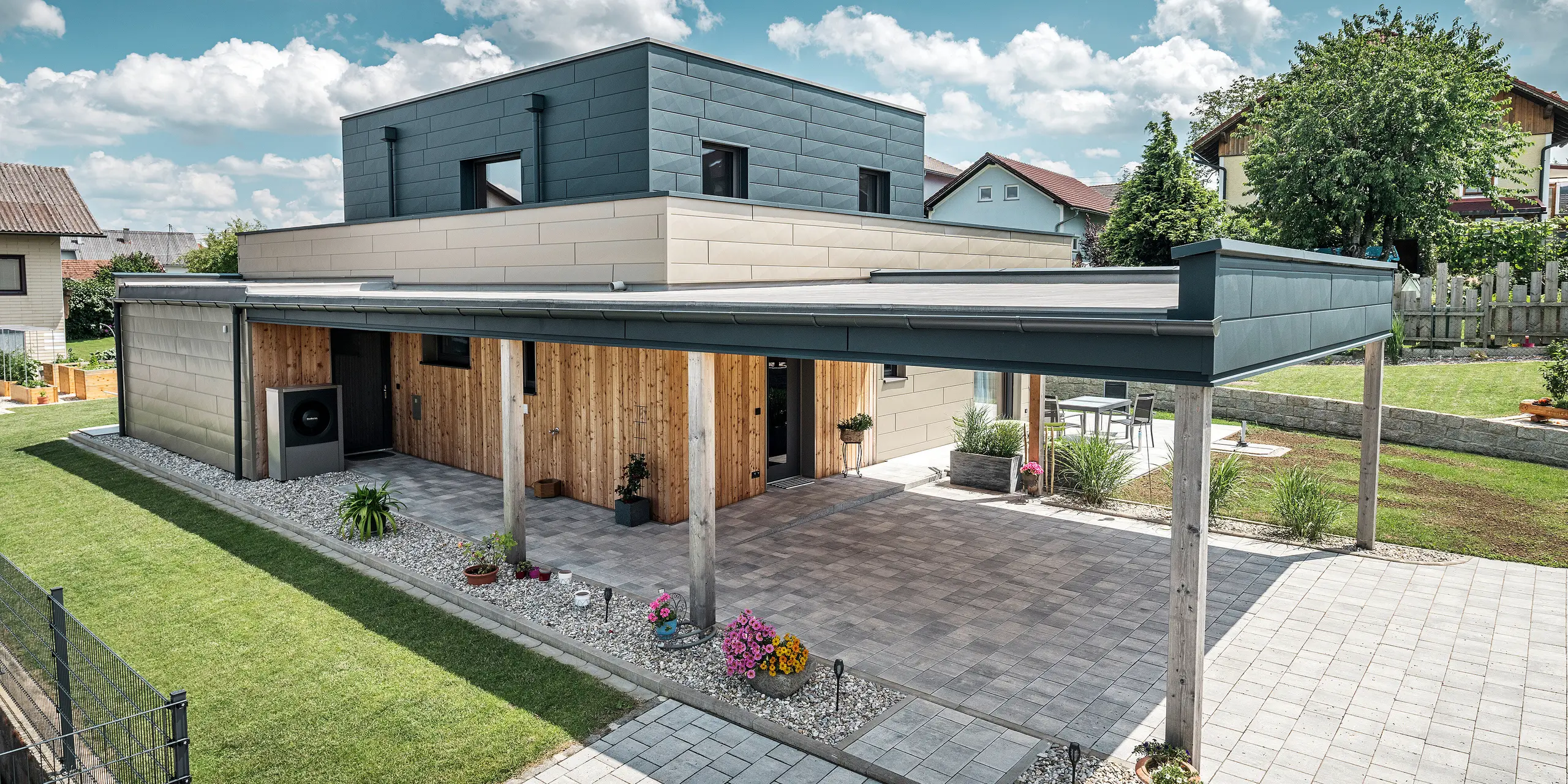Luftfoto af indkørslen til et moderne enfamiliehus i Wippenham, Østrig, med en tofarvet PREFA aluminiumsfacade i bronze og antracit. Huset viser den stilfulde integration af Siding.X facadepaneler og træbeklædning. Den overdækkede carport, der er integreret i husets arkitektur, er tydeligt synlig. Den velplejede have og terrasseområdet understreger bygningens moderne og funktionelle design.