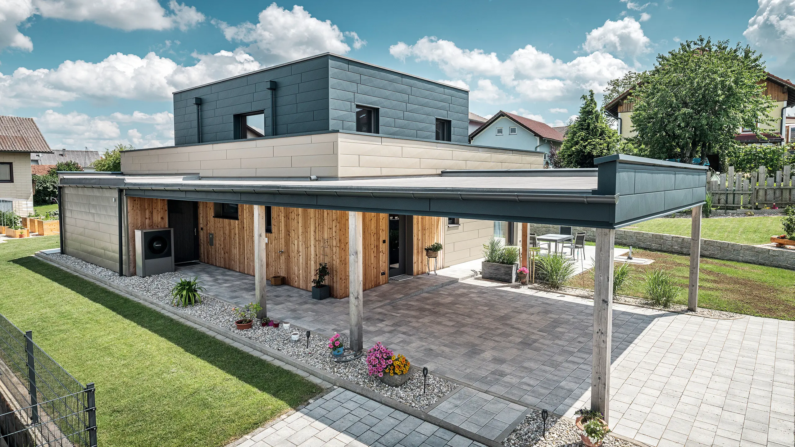 Luftfoto af indkørslen til et moderne enfamiliehus i Wippenham, Østrig, med en tofarvet PREFA aluminiumsfacade i bronze og antracit. Huset viser den stilfulde integration af Siding.X facadepaneler og træbeklædning. Den overdækkede carport, der er integreret i husets arkitektur, er tydeligt synlig. Den velplejede have og terrasseområdet understreger bygningens moderne og funktionelle design.