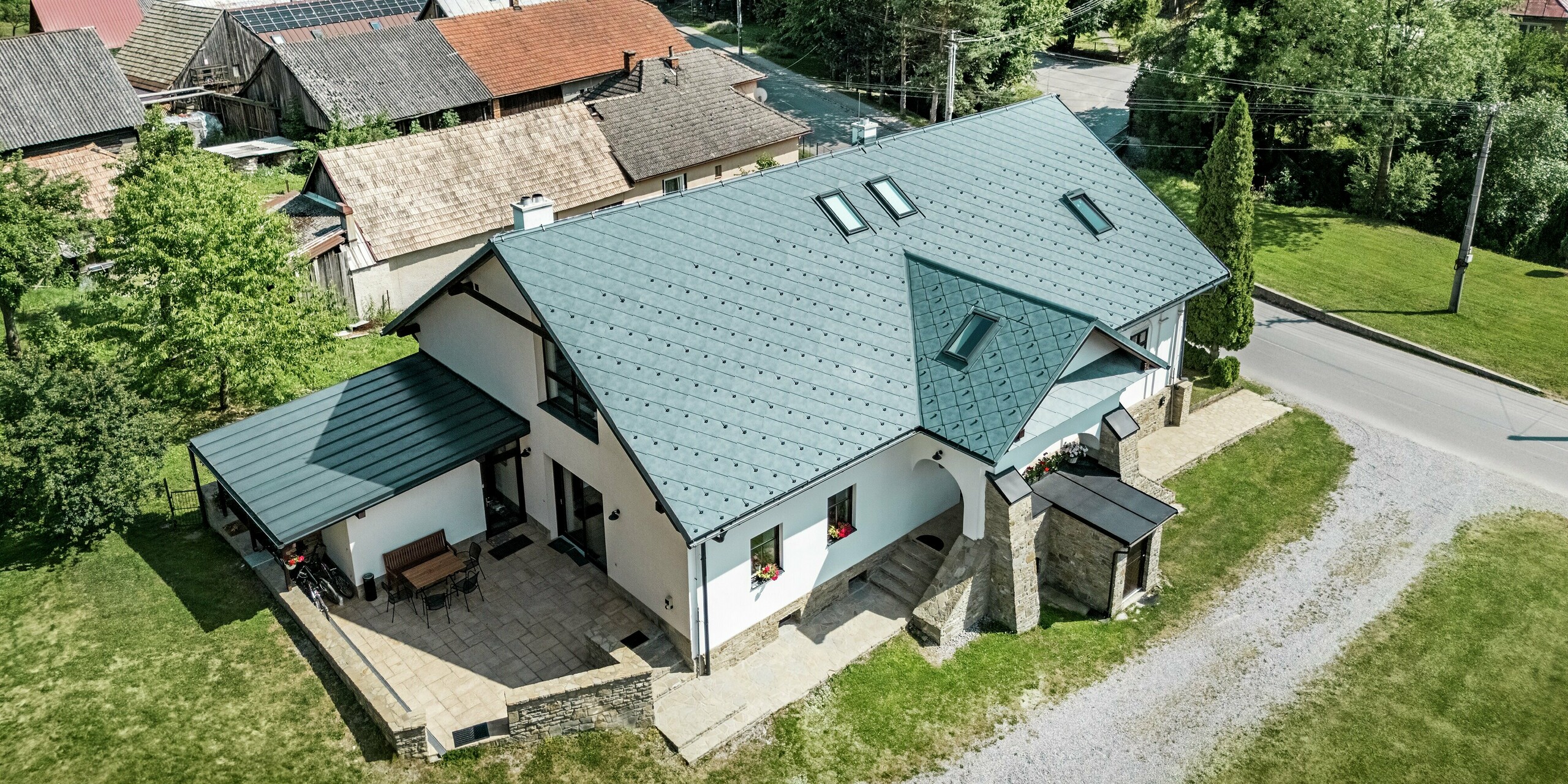 Das Bild zeigt das Handwerkerhaus in Klin, Slowakei, von oben. Das Satteldach, bedeckt mit PREFA Dachraute 44 × 44 in P.10 Steingrau, erstreckt sich elegant über das Gebäude und fügt sich harmonisch in die Umgebung ein. Die symmetrische Anordnung der Dachflächen und die integrierten Dachfenster sorgen für eine gleichmäßige Belichtung des Innenraums. Der moderne Stil des Daches kontrastiert perfekt mit der traditionellen Architektur des Hauses, das durch Natursteinmauerwerk und weiße Fassaden besticht. Das Aluminiumdach ist nicht nur ein ästhetisches Highlight, sondern auch äußerst funktional: Es ist leicht, langlebig, korrosionsbeständig und ideal für die Sanierung von historischen Gebäuden.
