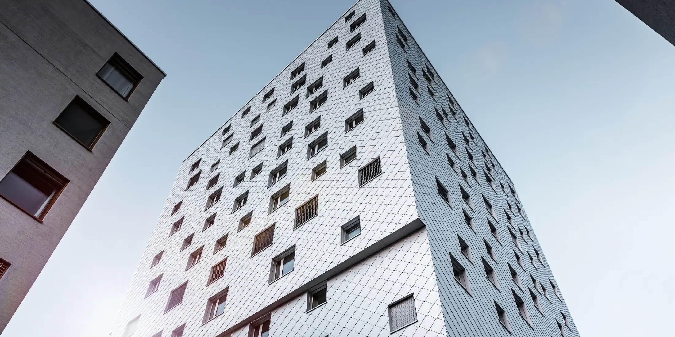 Das Bild zeigt eine Perspektive von unten auf das Solitaire-Hochhaus in Horw, Luzern, das von der Architektin Tilla Theus entworfen wurde. Die Fassade des Gebäudes ist mit eloxierten Aluminiumrauten von PREFA verkleidet, die ein diagonales Muster bilden und die Umgebung reflektieren. Die Fenster sind unregelmäßig angeordnet, was zu der charakteristischen Optik des Gebäudes beiträgt. Im Hintergrund sind Teile anderer Gebäude sichtbar, und der Himmel ist klar und blau.