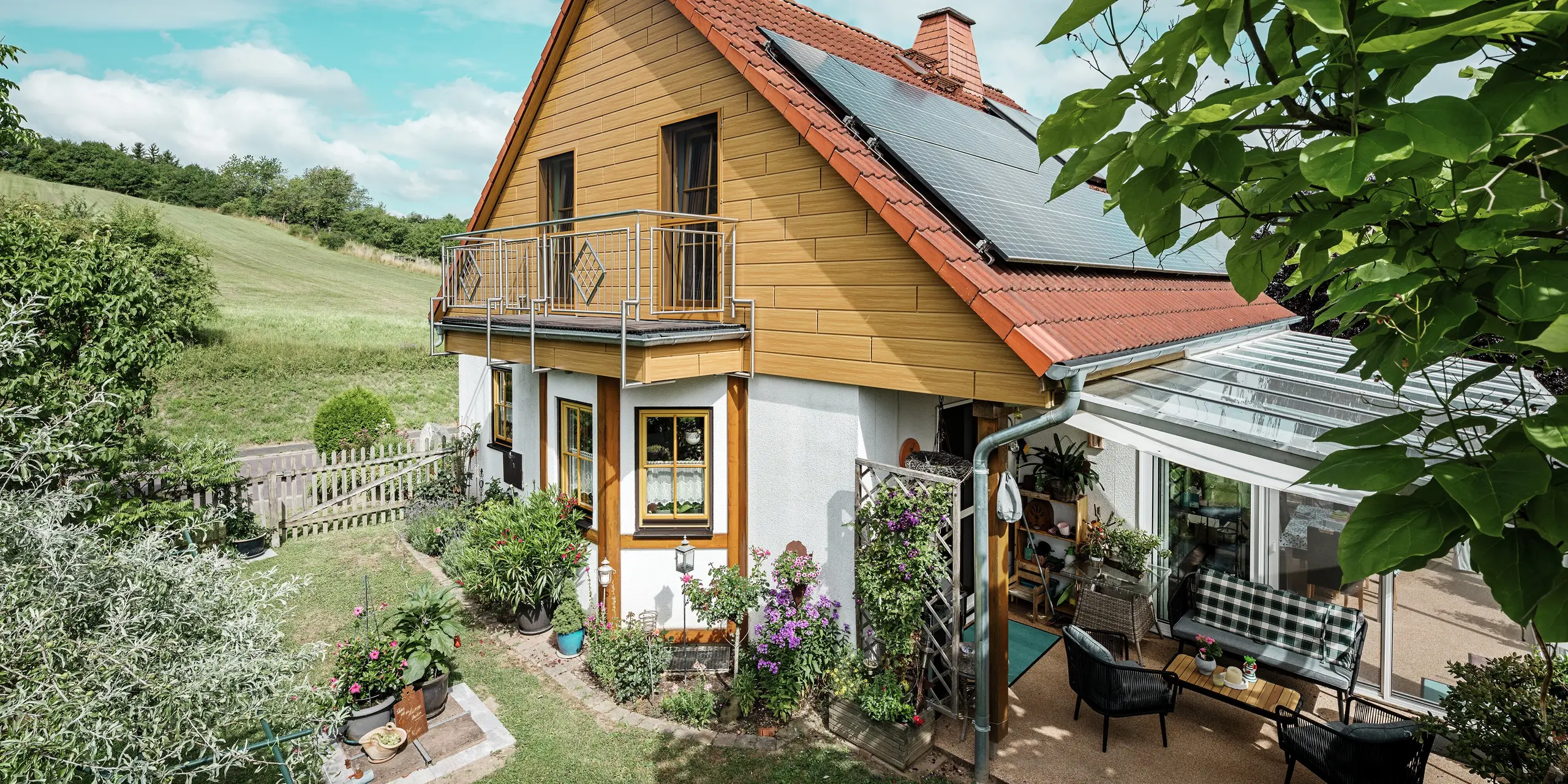 I dette billede vises et charmerende enfamilieshus i Blieskastel, hvis nyrenoverede gavl med PREFA Sidings i natureg straks falder i øjnene. Det varme trædesign af langtidsholdbart aluminium harmonerer imponerende med det røde tegltag og den store vinterhave. Disse omgivelser af yppigt grønt og omsorgsfuldt anlagte blomsterbede skaber en idyllisk og livfuld scene. Den robuste facaderenovering med PREFA giver ikke bare huset et naturligt udseende, men overbeviser også med dens vejrbestandighed, farveægthed og næsten vedligeholdelsesfri lange holdbarhed.