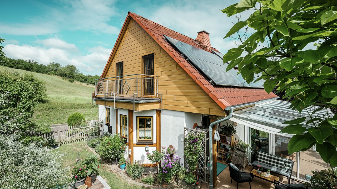 I dette billede vises et charmerende enfamilieshus i Blieskastel, hvis nyrenoverede gavl med PREFA Sidings i natureg straks falder i øjnene. Det varme trædesign af langtidsholdbart aluminium harmonerer imponerende med det røde tegltag og den store vinterhave. Disse omgivelser af yppigt grønt og omsorgsfuldt anlagte blomsterbede skaber en idyllisk og livfuld scene. Den robuste facaderenovering med PREFA giver ikke bare huset et naturligt udseende, men overbeviser også med dens vejrbestandighed, farveægthed og næsten vedligeholdelsesfri lange holdbarhed.