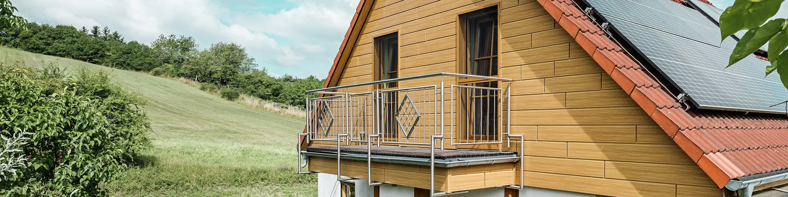 I dette billede vises et charmerende enfamilieshus i Blieskastel, hvis nyrenoverede gavl med PREFA Sidings i natureg straks falder i øjnene. Det varme trædesign af langtidsholdbart aluminium harmonerer imponerende med det røde tegltag og den store vinterhave. Disse omgivelser af yppigt grønt og omsorgsfuldt anlagte blomsterbede skaber en idyllisk og livfuld scene. Den robuste facaderenovering med PREFA giver ikke bare huset et naturligt udseende, men overbeviser også med dens vejrbestandighed, farveægthed og næsten vedligeholdelsesfri lange holdbarhed.