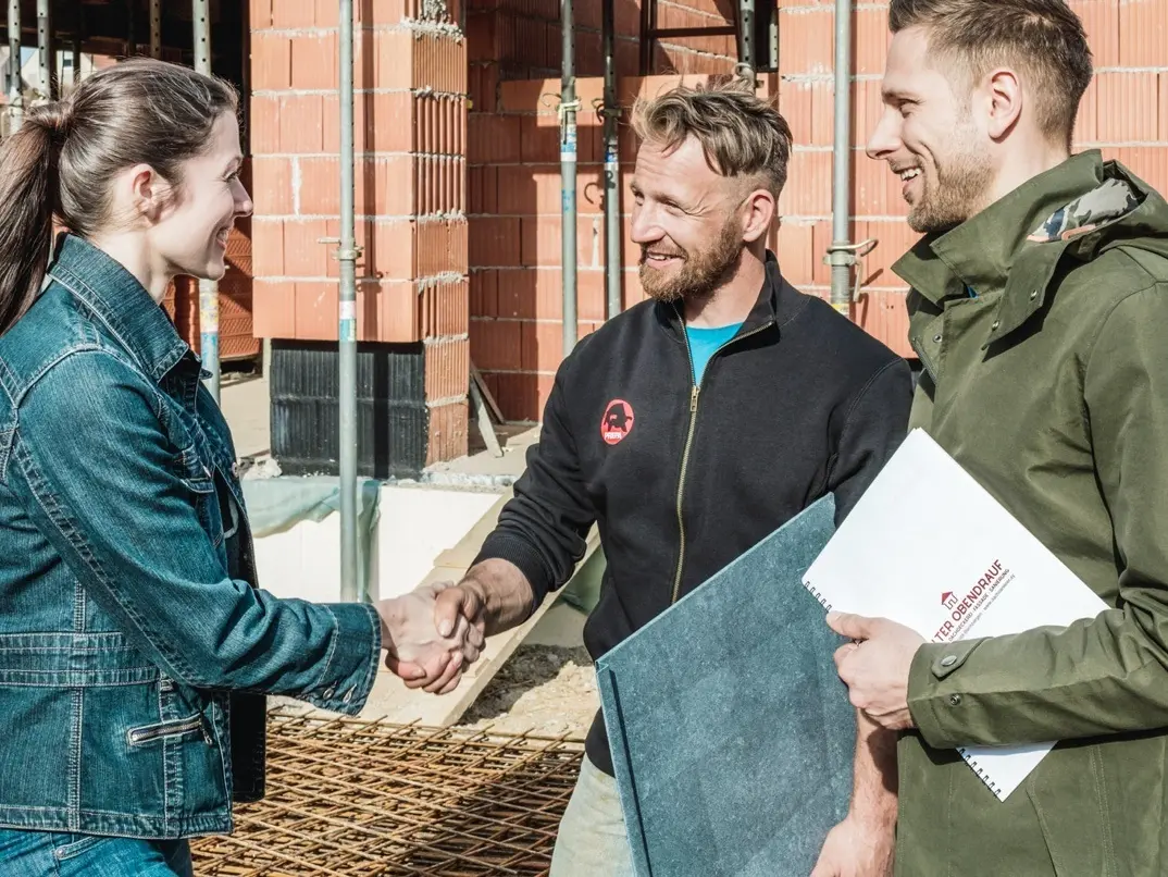 Et ungt par møder nabohåndværkeren på deres byggeplads. De har diskuteret tilbuddet og accepteret det. Blikkenslageren holder et PREFA-tagpanel i hånden.