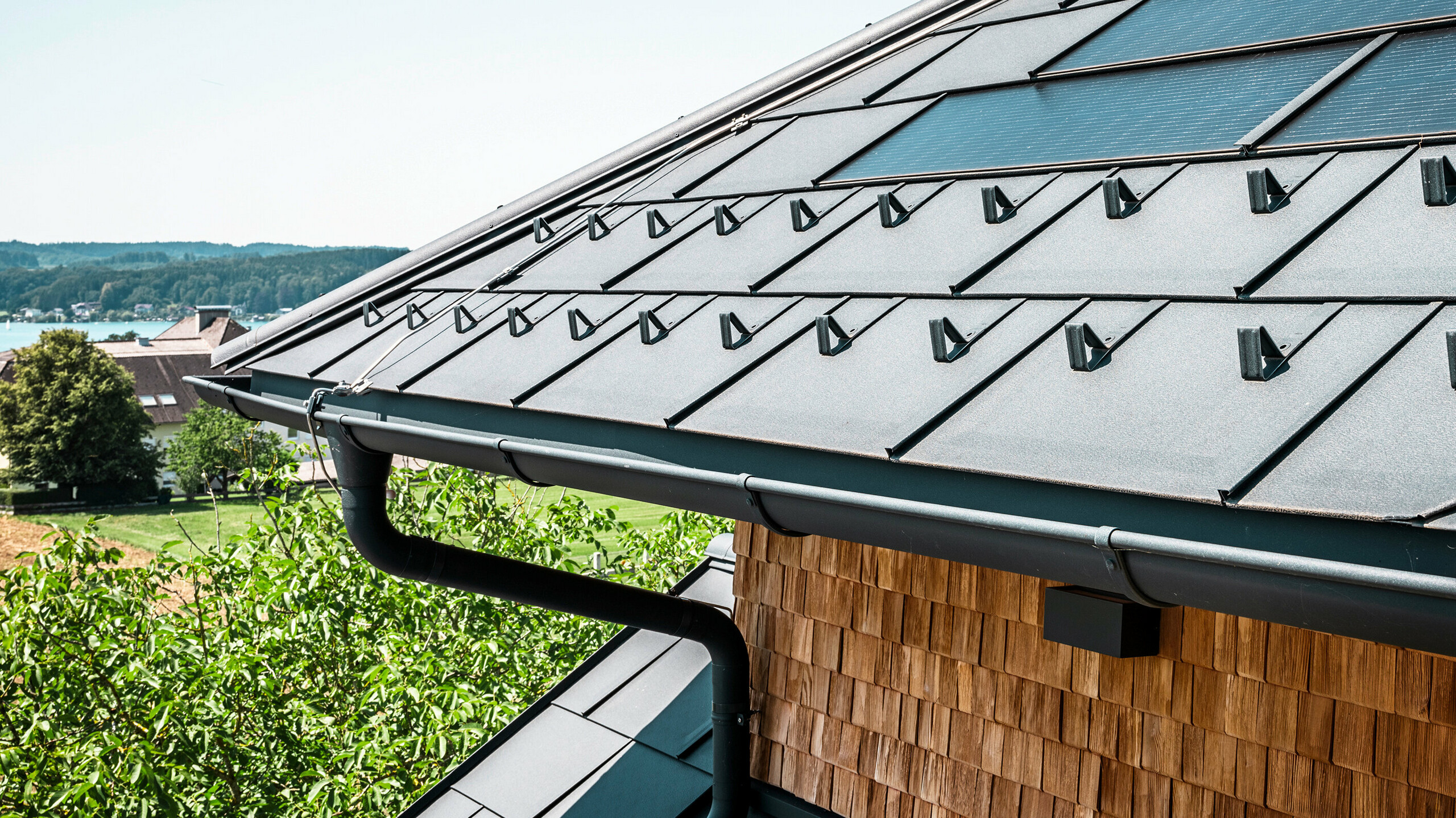 Zenz'n Stub'n i Schörfling ved Attersee har fået et nyrenoveret tag med innovative PREFA-solcelletagplader og holdbare PREFA-tagplader R.16 i P.10 antracit. Det stilfulde aluminiumstag kombinerer moderne design med bæredygtig energiproduktion. Tagafvandingen, der består af nedløbsrør og halvrunde tagrender i P.10 antracit, passer harmonisk ind i helheden og understreger den høje kvalitet. I baggrunden glimter Attersees blå vand.