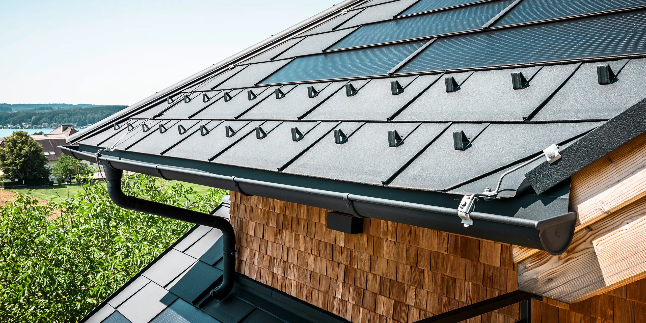 Zenz'n Stub'n i Schörfling ved Attersee har fået et nyrenoveret tag med innovative PREFA-solcelletagplader og holdbare PREFA-tagplader R.16 i P.10 antracit. Det stilfulde aluminiumstag kombinerer moderne design med bæredygtig energiproduktion. Tagafvandingen, der består af nedløbsrør og halvrunde tagrender i P.10 antracit, passer harmonisk ind i helheden og understreger den høje kvalitet. I baggrunden glimter Attersees blå vand.
