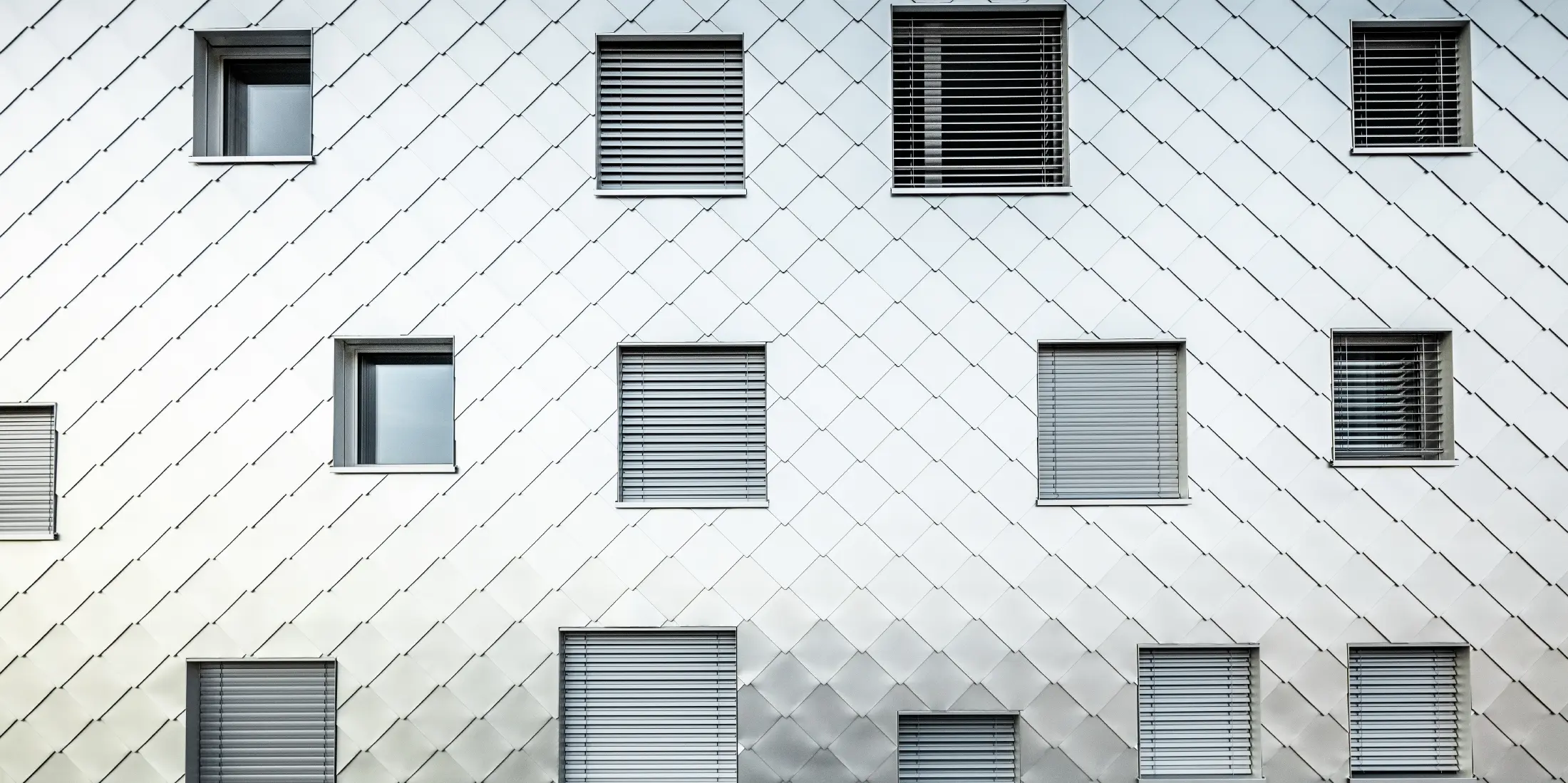 Das Bild zeigt eine Nahaufnahme der Fassade des Solitaire-Hochhauses in Horw, Luzern, das von der Architektin Tilla Theus entworfen wurde. Die Fassade ist mit eloxierten Aluminiumrauten verkleidet, die ein diagonales Muster bilden. Die Fenster sind unregelmäßig angeordnet und tragen zur einzigartigen Optik des Gebäudes bei. Einige Fenster sind mit Jalousien ausgestattet. Die Oberfläche der Aluminiumrauten reflektiert die Umgebung und verleiht der Fassade eine dynamische und glänzende Erscheinung.
