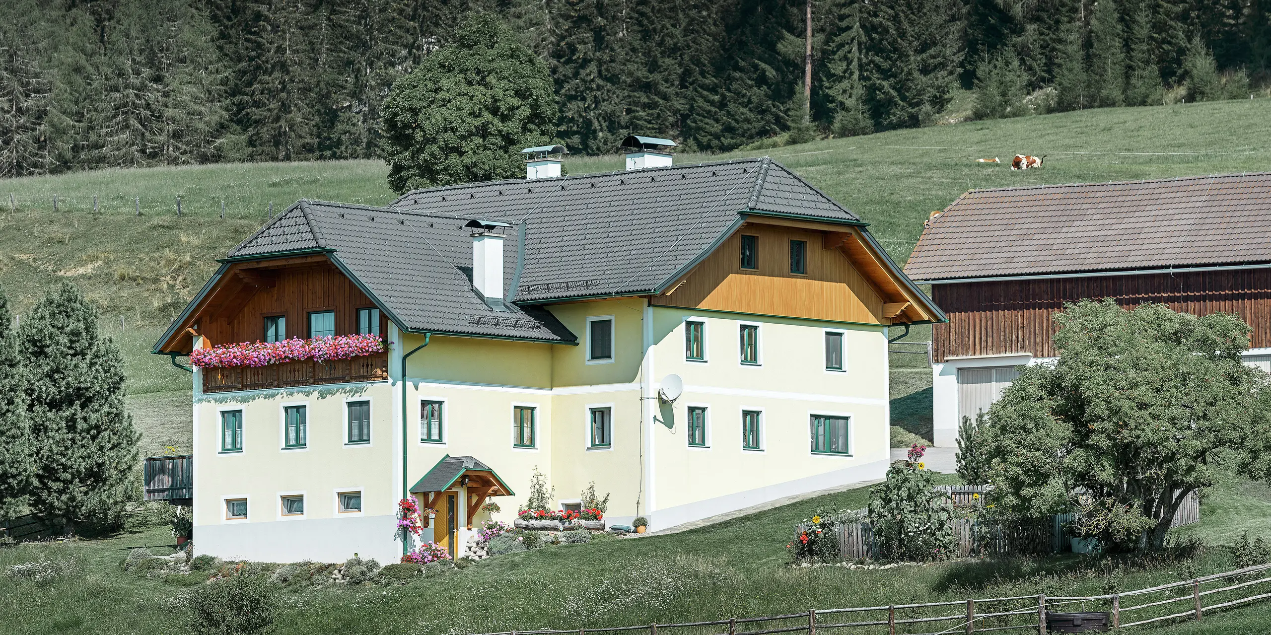 Panoramaaufnahme eines Landhauses in Ranten. Die Giebel des Hauses sind mit Holz und Aluminiumpaneelen in Holzoptik verkleidet. Der Unterschied zwischen Holz und Aluminium ist kaum zu erkennen. Die neu verkleidete Giebelseite wurde mit PREFA Sidings in Eiche natur ausgestattet. Das traditionelle Wohnhaus ist umgeben von Wiesen, Bäumen und einem angrenzenden Stadel.