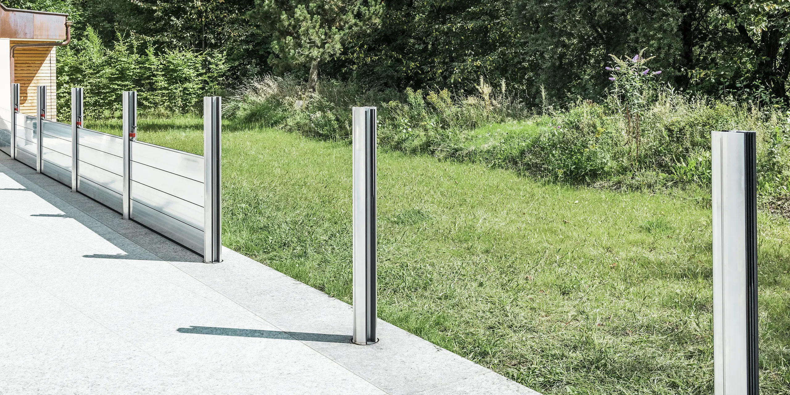 Detailansicht des mobilen PREFA Hochwasserschutzsystems beim Aufbau auf einer Terrasse in Tirol. Zu sehen sind bereits montierte Bodenhülsen sowie eingesetzte Aluminiumstützen, die das Dammbalkensystem aufnehmen. Im Hintergrund erkennt man die angrenzende Wiese und das offene Gelände - ein typisches Einsatzgebiet für den PREFA Landschaftsschutz. Die hochwertige Aluminiumkonstruktion bietet zuverlässigen Schutz vor Hochwasser bei eleganter Optik. In den beiden vorderen Stützen sind noch keine Dammbalken eingesetzt, da sich das System zum Zeitpunkt des Fotos in der Aufbauphase befindet.