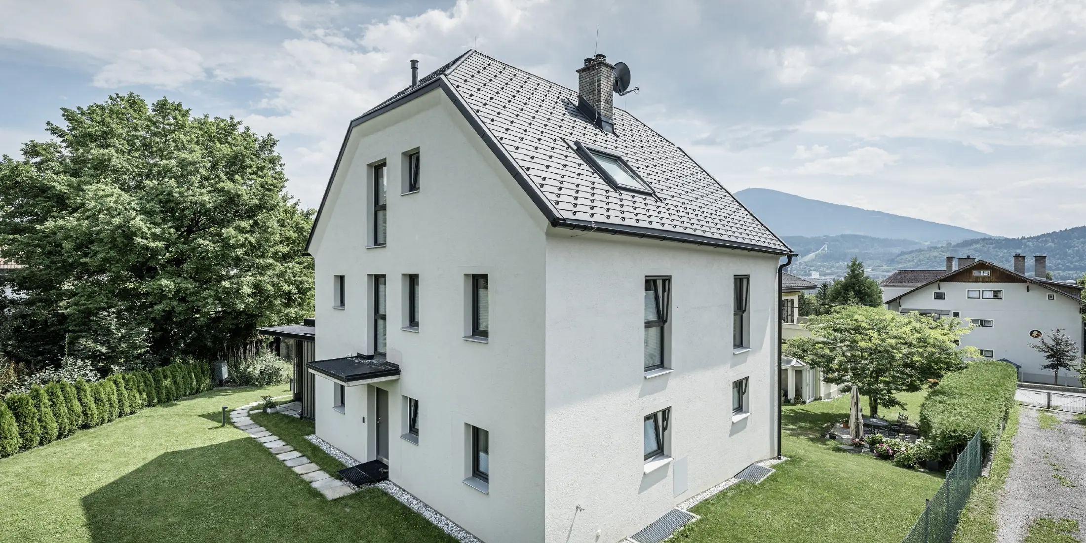 Moderne treetagers enfamiliehus med lys pudset facade og mørk grå sadeltag af aluminium fra PREFA. Bygningen har flere smalle vinduer og tagafvanding i antracit. Den ligger i en velplejet have med græsplæne, træer og hække, og i baggrunden ses flere boliger og et bjergrigt landskab.
