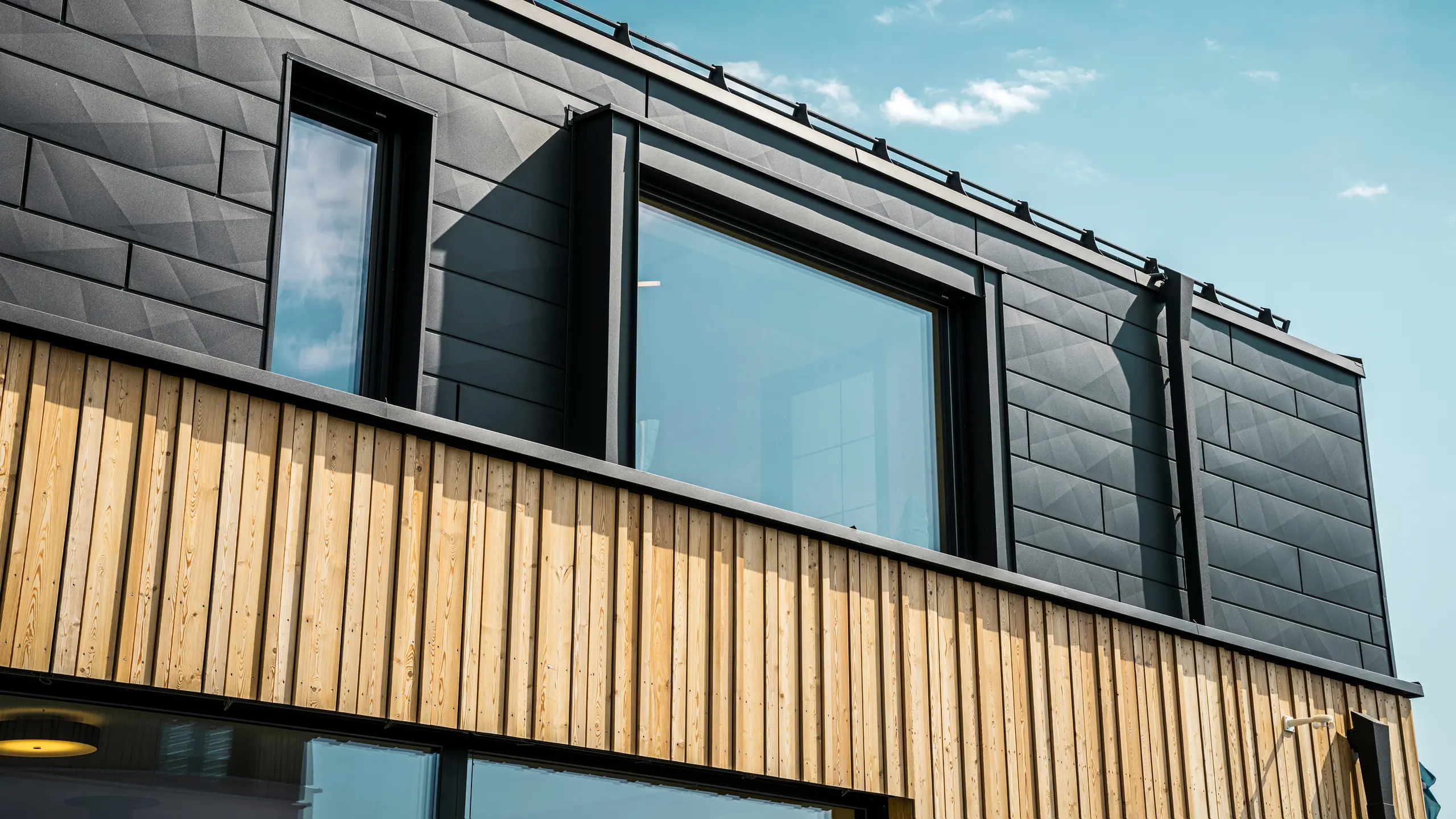 Nærbillede af øverste etage i modelhuset Autark i præfabrikationscenteret Blaue Lagune, Vösendorf (AT). Facaden består af PREFA Siding.X aluminiumpaneler i P.10 sort, der med deres specielle udseende med forskellige kantninger er meget iøjnefaldende. Derunder findes en stilfuld træbeklædning, der danner en harmonisk kontrast til det mørke aluminium. Store vinduer lukker meget lys ind i bygningen og understreger det moderne design.