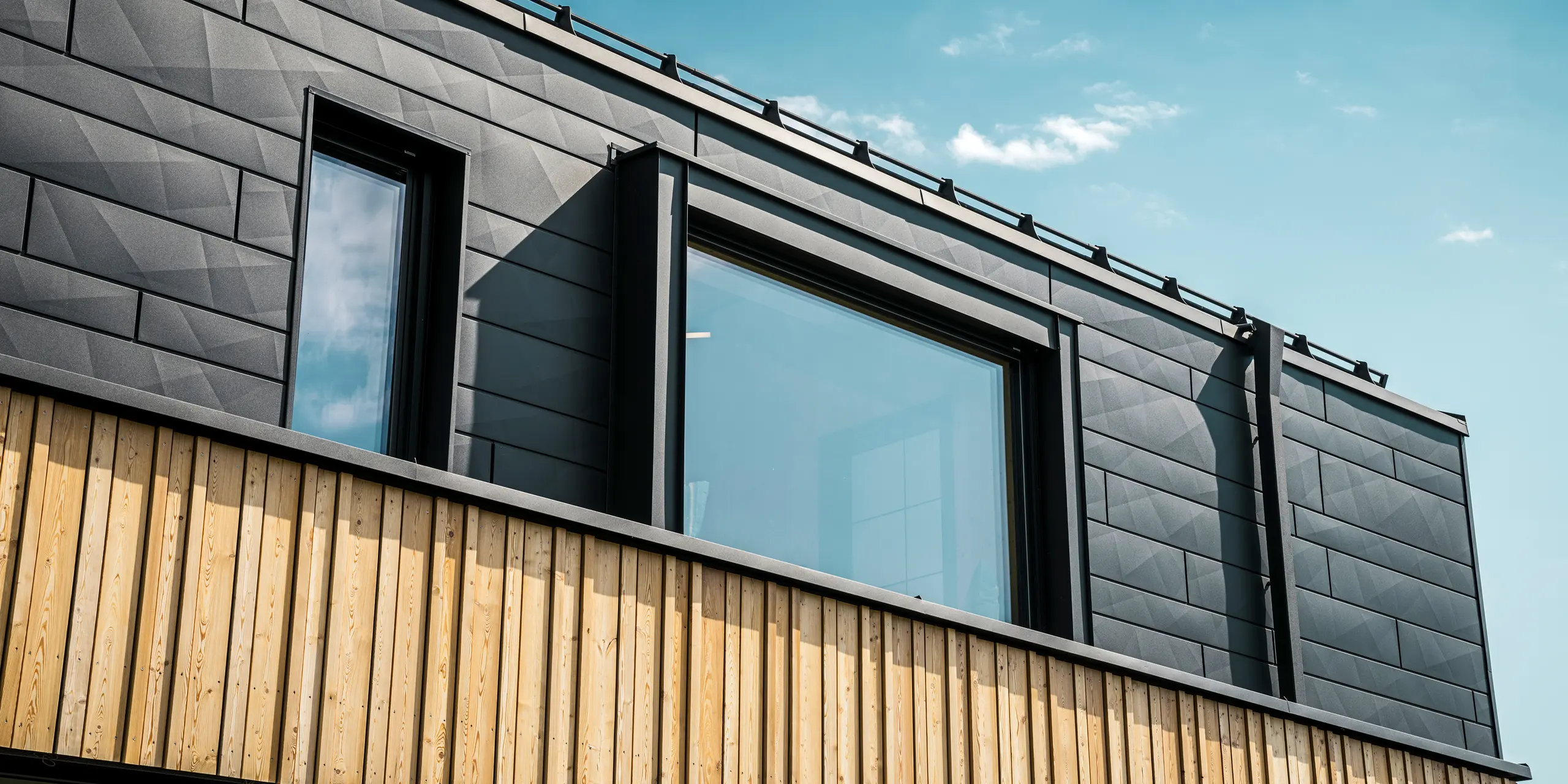 Nærbillede af øverste etage i modelhuset Autark i præfabrikationscenteret Blaue Lagune, Vösendorf (AT). Facaden består af PREFA Siding.X aluminiumpaneler i P.10 sort, der med deres specielle udseende med forskellige kantninger er meget iøjnefaldende. Derunder findes en stilfuld træbeklædning, der danner en harmonisk kontrast til det mørke aluminium. Store vinduer lukker meget lys ind i bygningen og understreger det moderne design.