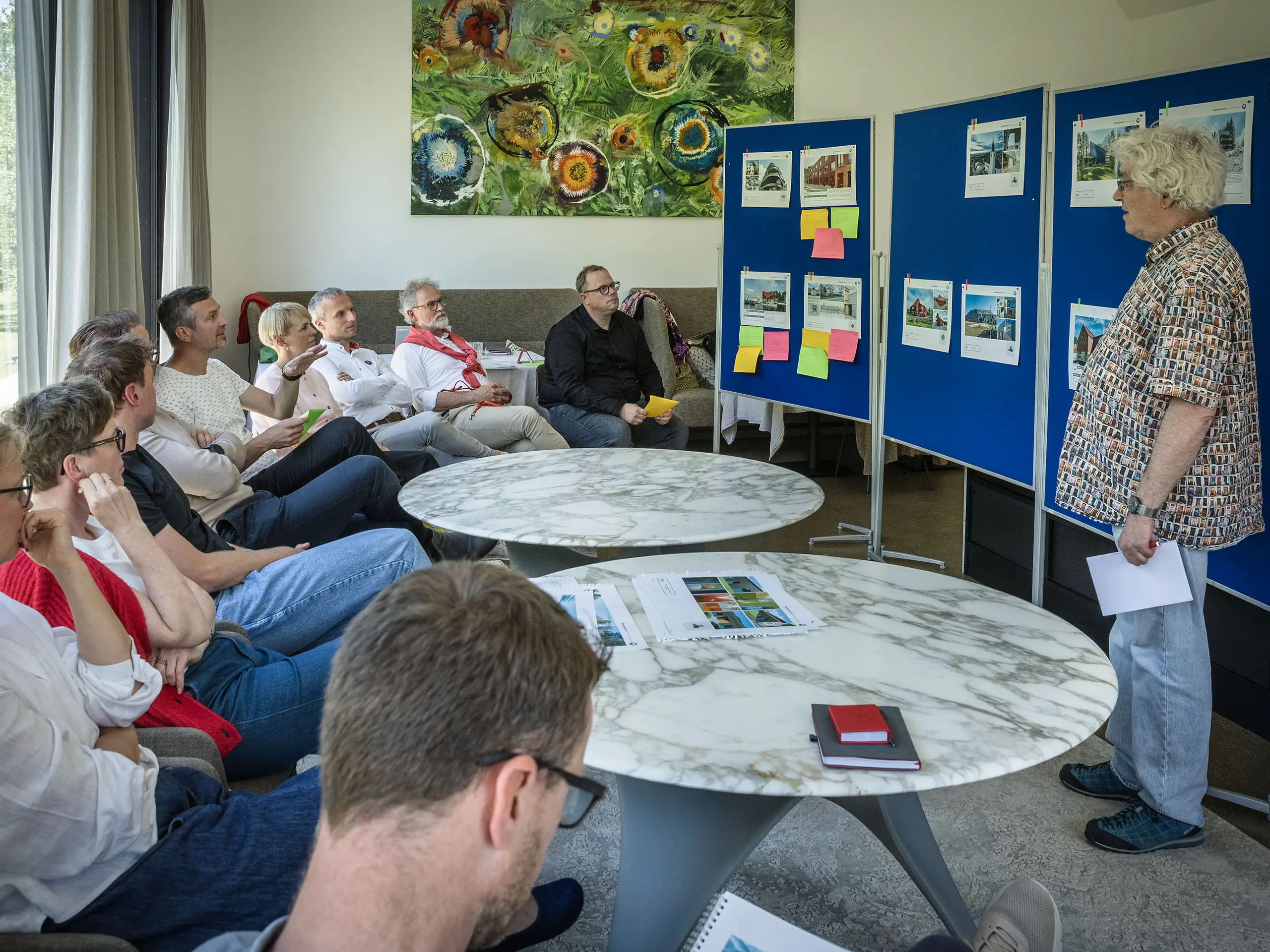 PREFARENZEN 2027 Jurysitzung in einem hellen Seminarraum: Moderator Matthias Finkentey präsentiert vor blauen Pinnwänden Architekturprojekte aus PREFA Aluminium, während die Jury aufmerksam sitzt und Notizen macht. Große Fenster lassen Tageslicht herein und geben den Blick auf einen grünen Garten frei. Auf den Tischen liegen Architekturbücher, Notizhefte und Stifte. Die Szene vermittelt konzentrierte Analyse, kreativen Austausch und Bewertung von Design und Funktion. PREFA Aluminiumprodukte symbolisieren langlebige, innovative Dächer und Fassaden, die Ästhetik, Qualität und Inspiration für Architekturprojekte vereinen.