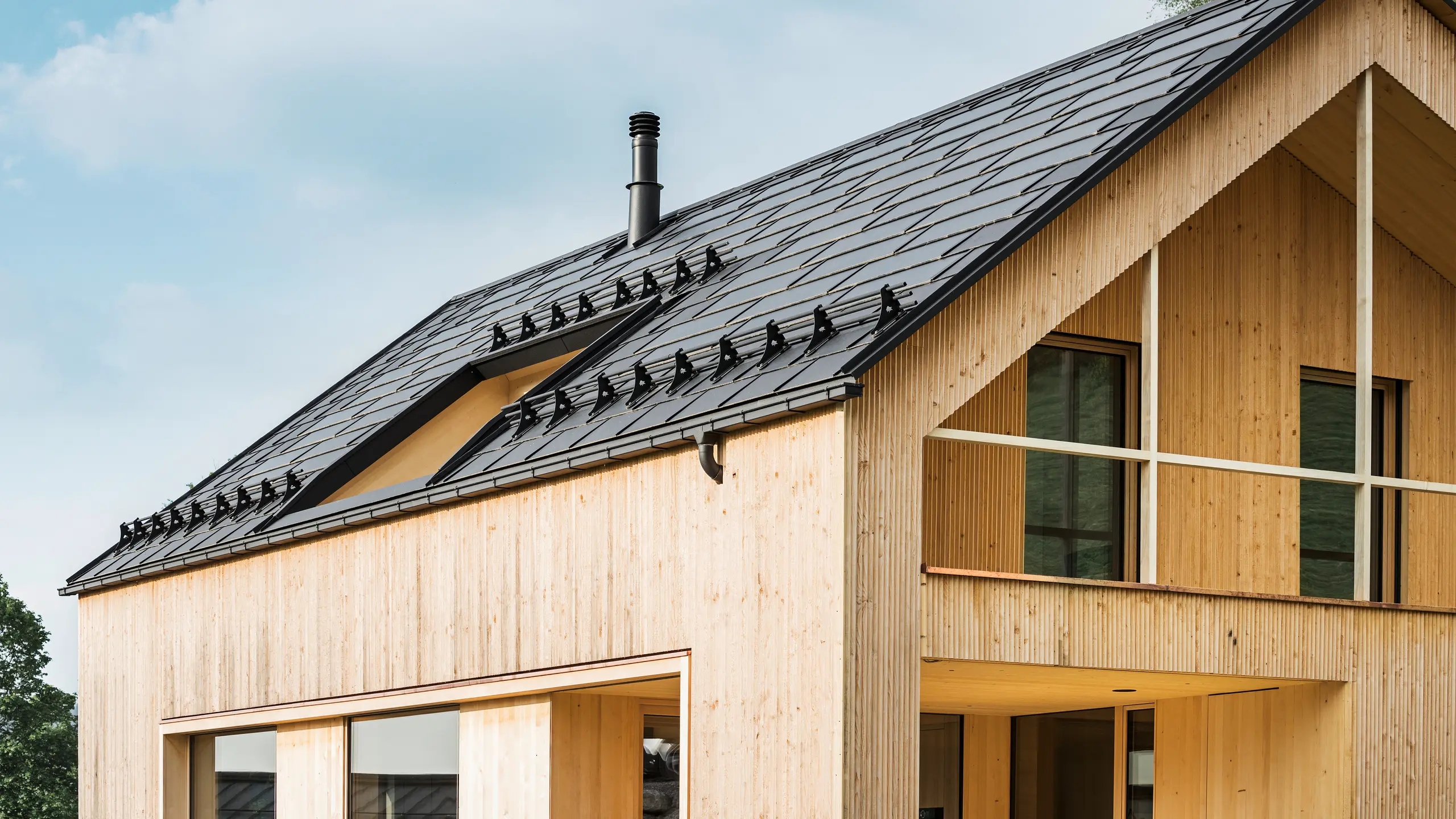 Nærbillede af gavlen på et miljøvenligt hus i Egg, der er kendetegnet ved PREFA-solcelletagplader i elegant sort, som harmonerer perfekt med PREFA-tagplader R.16. Tagets rene linjer står i kontrast til træfacadens naturlige struktur, mens solcellemodulerne er et statement for bæredygtigt boligbyggeri. Den diskrete skorsten passer ind i det samlede billede, der forener miljøbevidst design med landlig æstetik. Desuden har det innovative økohus to altaner, der giver udsigt til den fantastiske bjergkulisse i Egg i Vorarlberg.