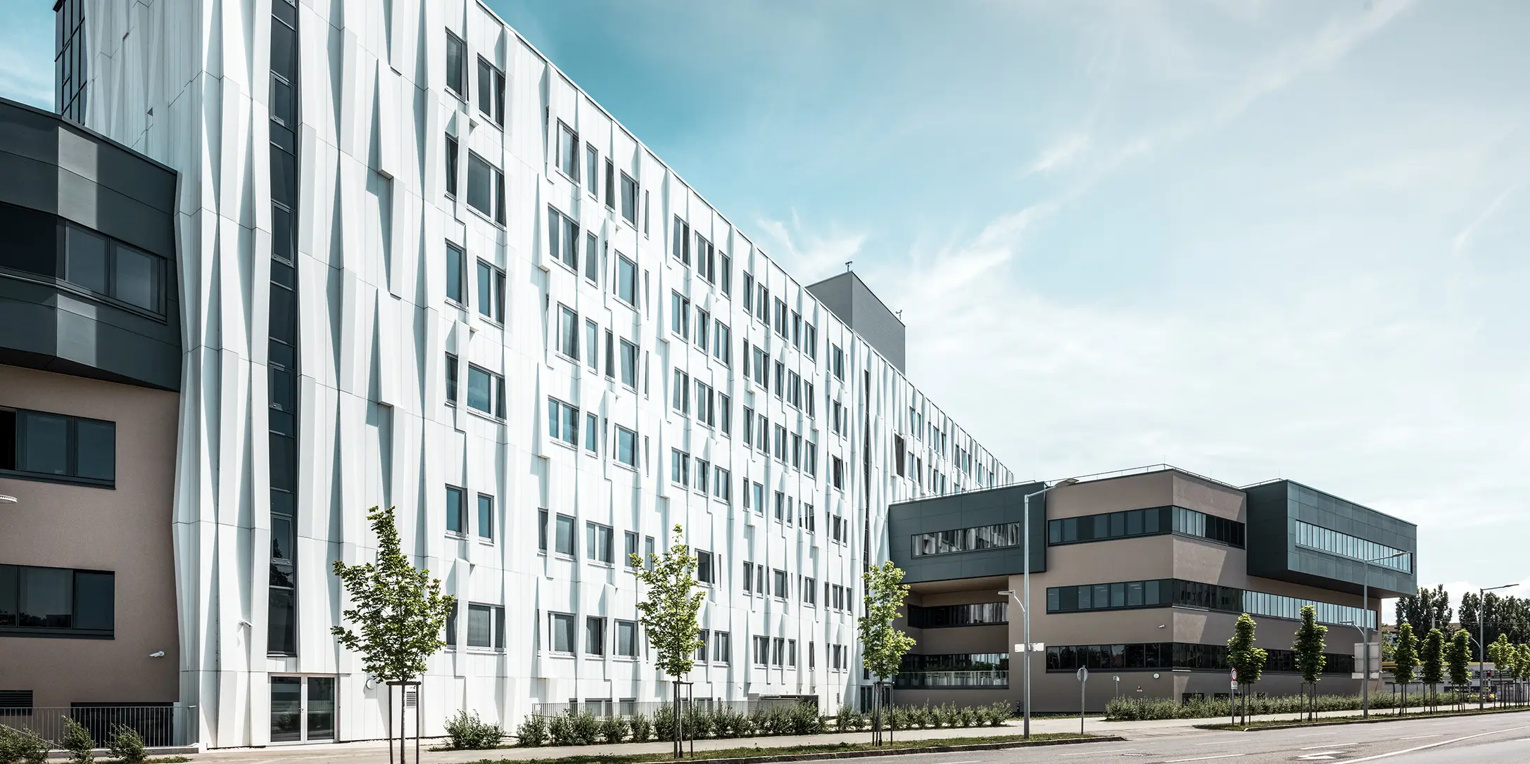 Sidebillede af universitetshospitalet i St. Pölten, Niederösterreich. Den robuste aluminiumsfacade gør bygningen til et blikfang i byen. I forgrunden ses en gade med nogle træer.