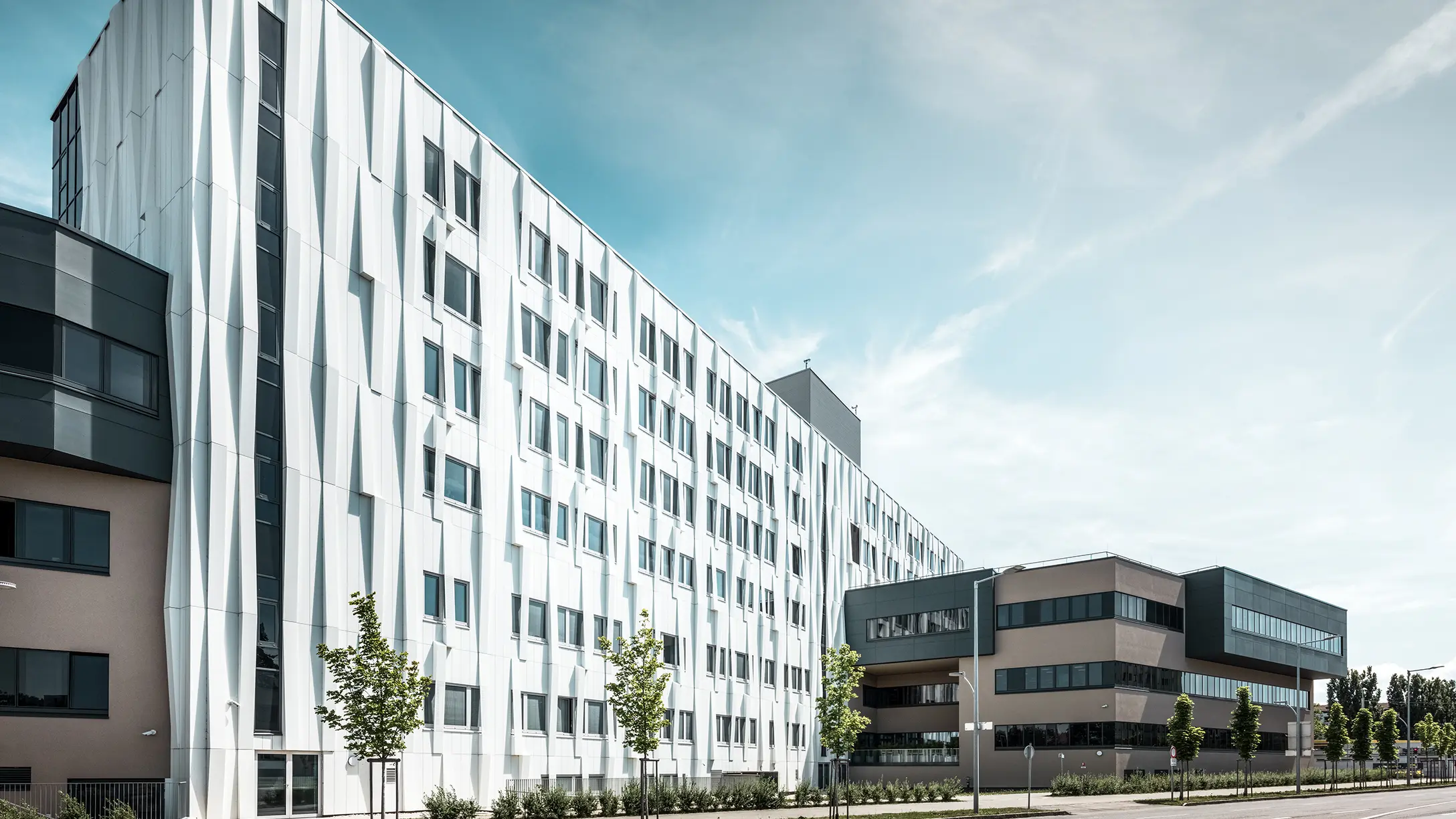 Sidebillede af universitetshospitalet i St. Pölten, Niederösterreich. Den robuste aluminiumsfacade gør bygningen til et blikfang i byen. I forgrunden ses en gade med nogle træer.