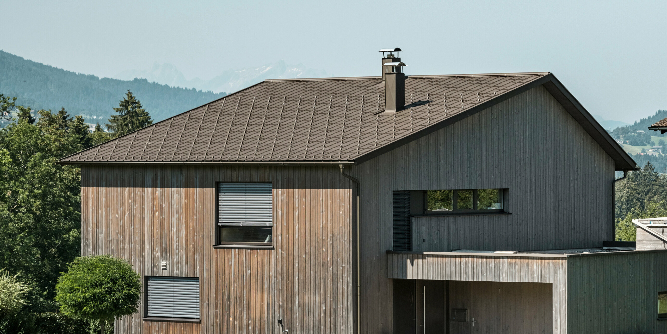 Sidebillede af et moderne parcelhus i Krumbach med en kombination af en naturlig træfacade og et holdbart PREFA-tag af aluminium i P.10 brun. Det harmoniske farveskema viser den elegante integration af moderne design og traditionelle materialer, hvor PREFA tagpladerne giver et æstetisk og funktionelt tagdesign.
