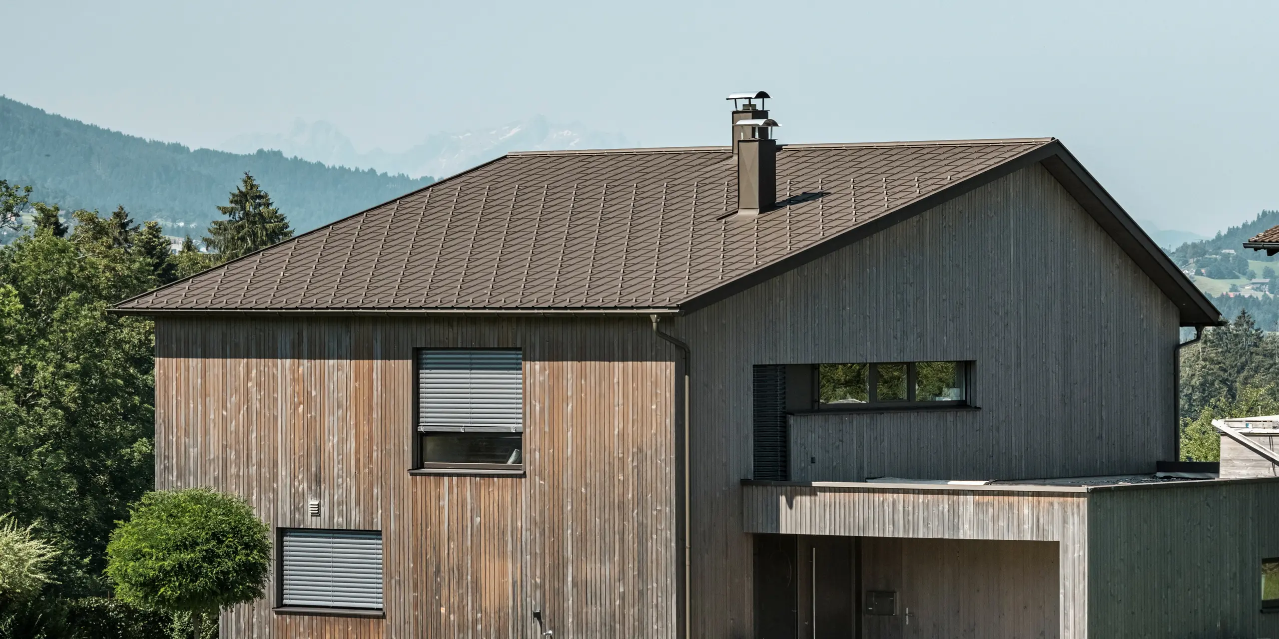 Sidebillede af et moderne parcelhus i Krumbach med en kombination af en naturlig træfacade og et holdbart PREFA-tag af aluminium i P.10 brun. Det harmoniske farveskema viser den elegante integration af moderne design og traditionelle materialer, hvor PREFA tagpladerne giver et æstetisk og funktionelt tagdesign.