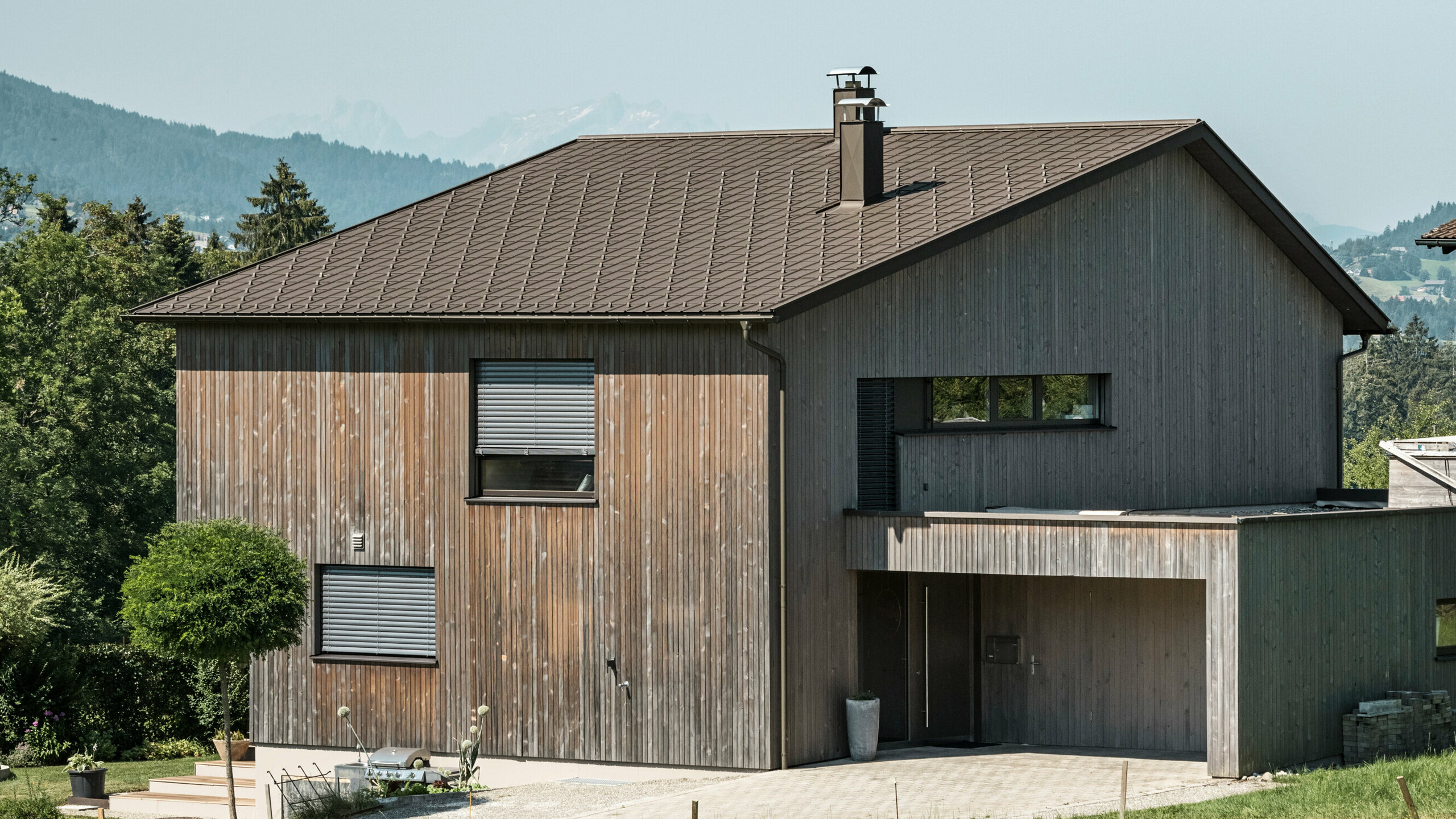 Sidebillede af et moderne parcelhus i Krumbach med en kombination af en naturlig træfacade og et holdbart PREFA-tag af aluminium i P.10 brun. Det harmoniske farveskema viser den elegante integration af moderne design og traditionelle materialer, hvor PREFA tagpladerne giver et æstetisk og funktionelt tagdesign.