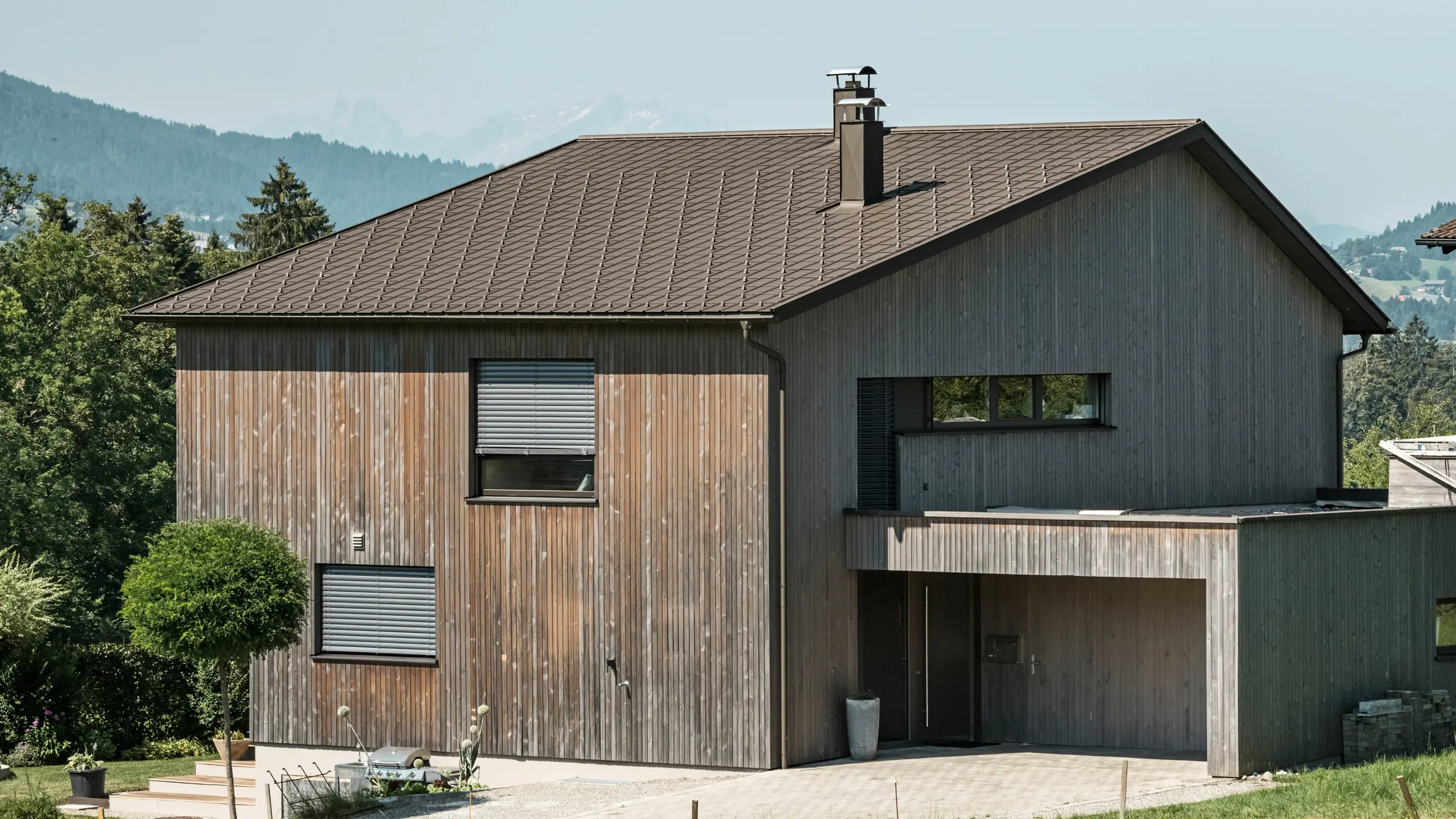 Sidebillede af et moderne parcelhus i Krumbach med en kombination af en naturlig træfacade og et holdbart PREFA-tag af aluminium i P.10 brun. Det harmoniske farveskema viser den elegante integration af moderne design og traditionelle materialer, hvor PREFA tagpladerne giver et æstetisk og funktionelt tagdesign.
