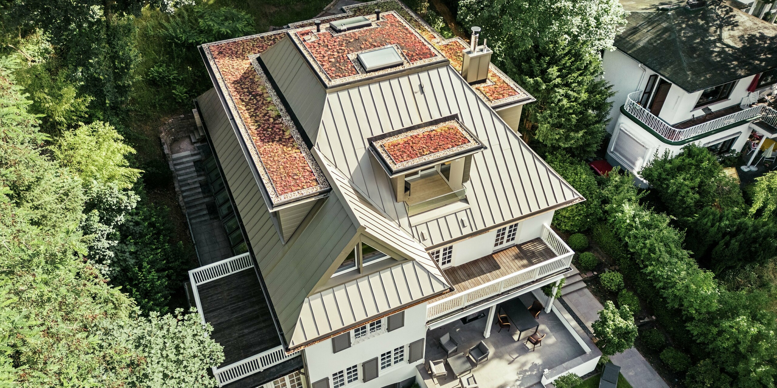 Die Luftaufnahme zeigt die Villa Blankenese in Hamburg mit ihrem markanten Dach aus PREFALZ in der Farbe Bronze. Die komplexe Dachform mit mehreren Gauben und integrierten Flächen wird durch die elegante Stehfalztechnik betont. Ergänzt wird das Design durch bronzefarbene PREFA Sidings an den Fassadenteilen, die für eine harmonische Verbindung von Dach und Fassade sorgen. Die Kombination aus langlebigem Aluminium und edler Farbgebung macht das Gebäude zu einem stilvollen und wetterfesten Blickfang inmitten grüner Umgebung.