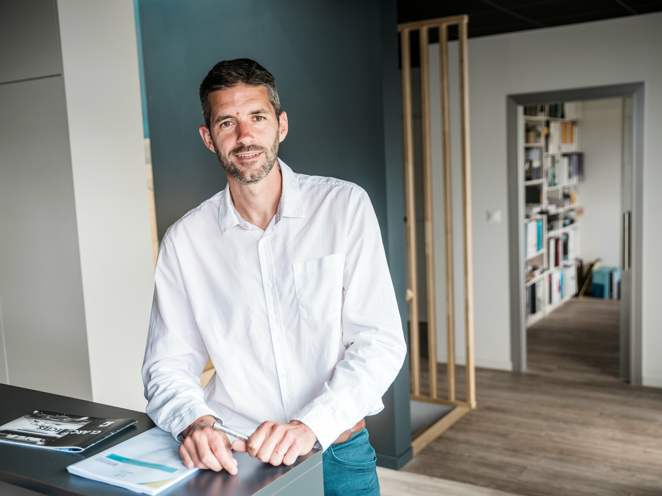 Im Vordergrund des Bildes steht Architekt Hugues Launay vom Architekturbüro CLARC Architectes. Der Mann ist zentral bzw. leicht links platziert und trägt formelle Kleidung, passend für ein professionelles Interview. Der Hintergrund zeigt die geschwungene Aluminiumfassade des Collège Léontine Dolivet, deren silbermetallic Oberfläche Lichtreflexe erzeugt. Die Person blickt leicht seitlich, die Körperhaltung wirkt präsent und selbstbewusst, was auf eine Rolle als Projektverantwortlicher schließen lässt. Die Szene ist bei Tageslicht aufgenommen, mit weichem, natürlichem Licht, das die Konturen klar hervorhebt.