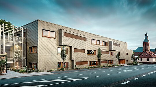 Die Aufnahme aus Straßenniveau zeigt die neue Kindertagesstätte in Alzenau im spannungsvollen Dialog mit der historischen Stadtkulisse, darunter die markante Kirche im Hintergrund. Die moderne Fassade aus bronzefarbenen PREFA Wandrauten 44 × 44 verleiht dem Gebäude eine elegante, fast skulpturale Wirkung. Präzise gesetzte Fenster, vertikale Holzlamellen und versetzte Fassadenflächen schaffen Tiefe und Rhythmus. Links ist das filigrane Treppenhaus mit weißem Metallgerüst sichtbar, das moderne Leichtigkeit vermittelt. Die klare Formsprache, nachhaltige Materialwahl und die hochwertige Aluminiumfassade machen das Gebäude zu einem Vorzeigeprojekt für städtebauliche Integration und zukunftsorientiertes Bauen mit PREFA Systemen.