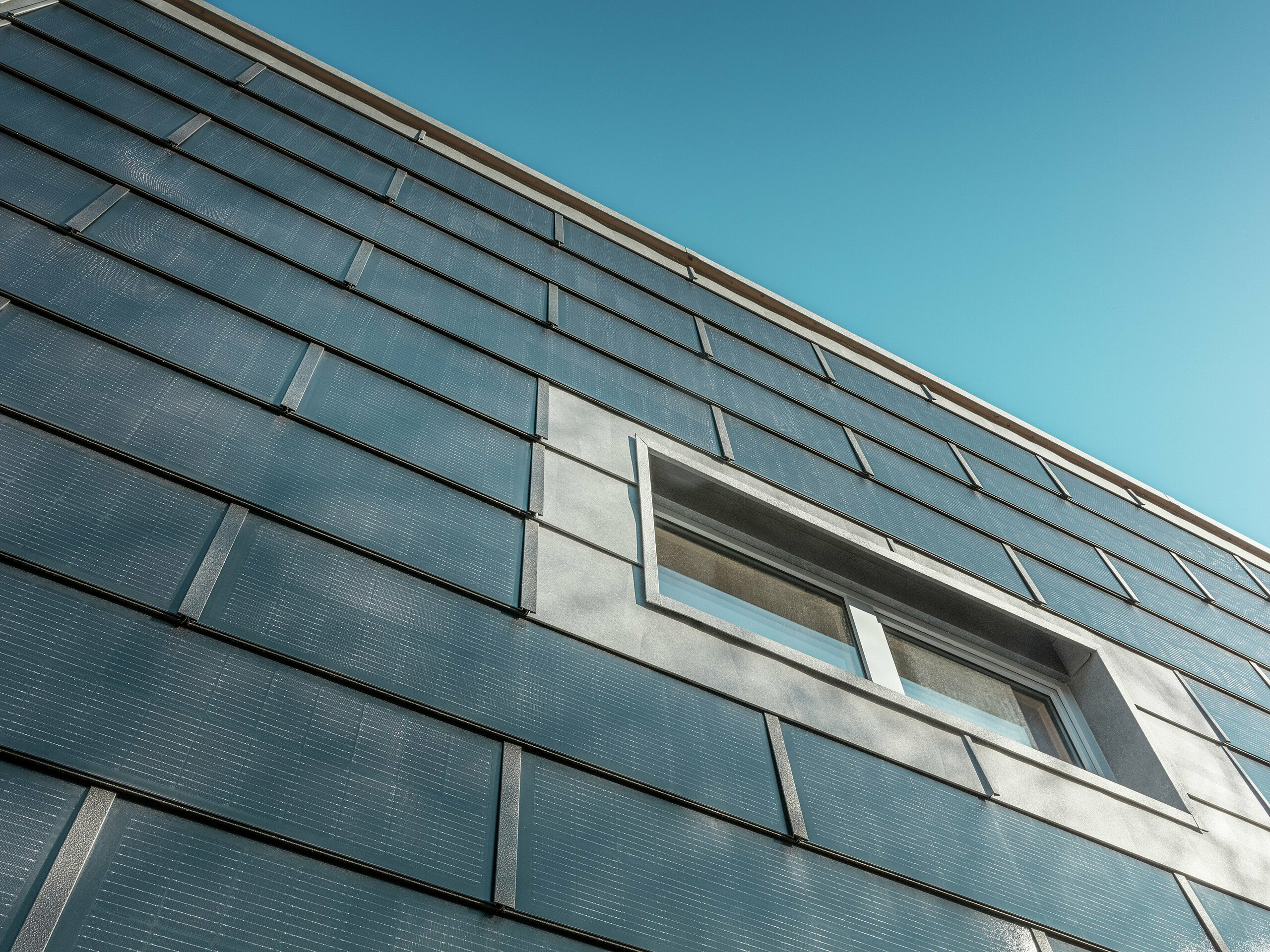 Die Fassade der RÜMA-Blechwerkstatt in Vorarlberg, aus einem bestimmten Winkel fotografiert, ist mit PREFA-Solardachziegeln verkleidet. Graue Aluminiumelemente und integrierte Photovoltaikmodule rahmen das Fenster ein. Im Hintergrund ist der klare blaue Himmel zu sehen.
