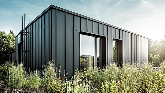 Die Aufnahme zeigt die moderne, kubische Dachterrasse der Budapester Mehrgenerationen-Villa, verkleidet mit hochwertigem PREFALZ Aluminium in P.10 Anthrazit. Die klar strukturierten Winkelstehfalze erzeugen eine elegante, vertikale Linienführung, die durch das weiche Sonnenlicht besonders betont wird. Umgeben von dichter, natürlicher Bepflanzung entsteht ein spannender Kontrast zwischen moderner Metallarchitektur und üppigem Grün. Die langlebigen, witterungsbeständigen PREFA Aluminiumprodukte unterstreichen die hochwertige Ausführung und machen die Dachterrasse zu einem markanten, architektonischen Highlight.