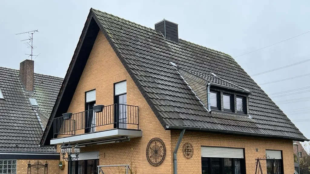 Enkeltfamiliehus af gul klinker med stejlt sadeltag og mørk tagdækning før tagrenoveringen; på havsiden med altan, tagkvist, terrasse med siddepladser og græsplæne i forgrunden.