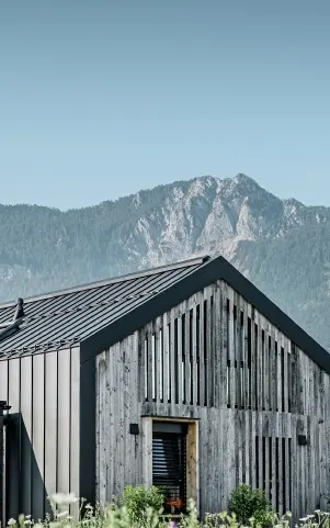 Moderner, funktionaler Wohnbau mit einer Kombination aus Holzfassade und PREFA Band- und Blechmaterial für das Dach. Die klare Architektur wird durch die elegante, metallische Dach- und Fassadenverkleidung unterstrichen, die ideal für anspruchsvolle Projekte ist. Das Bild veranschaulicht die vielseitigen Anwendungsmöglichkeiten der PREFA Produkte im Bereich Bänder und Bleche.