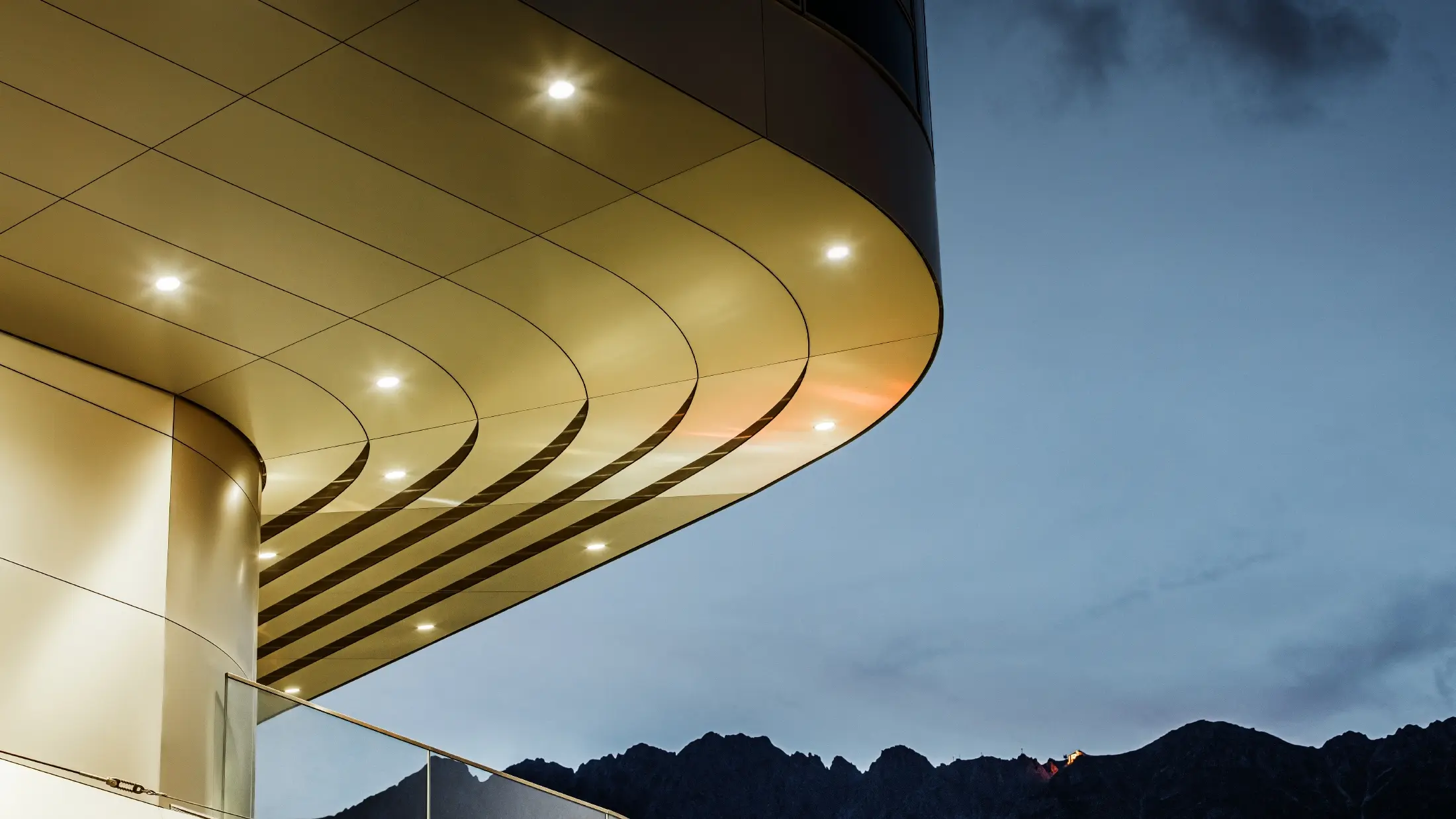 Billedet viser en imponerende arkitektonisk detalje af en PREFA PEMA-kompositplade af børstet aluminium. Pladen er let buet og har elegante, flydende linjer, der fremhæves særligt om aftenen ved hjælp af målrettet belysning. Pladens klare struktur og elegante aluminiumlook supplerer facadeens moderne design og tilbyder en elegant, moderne løsning til arkitektur af høj kvalitet.