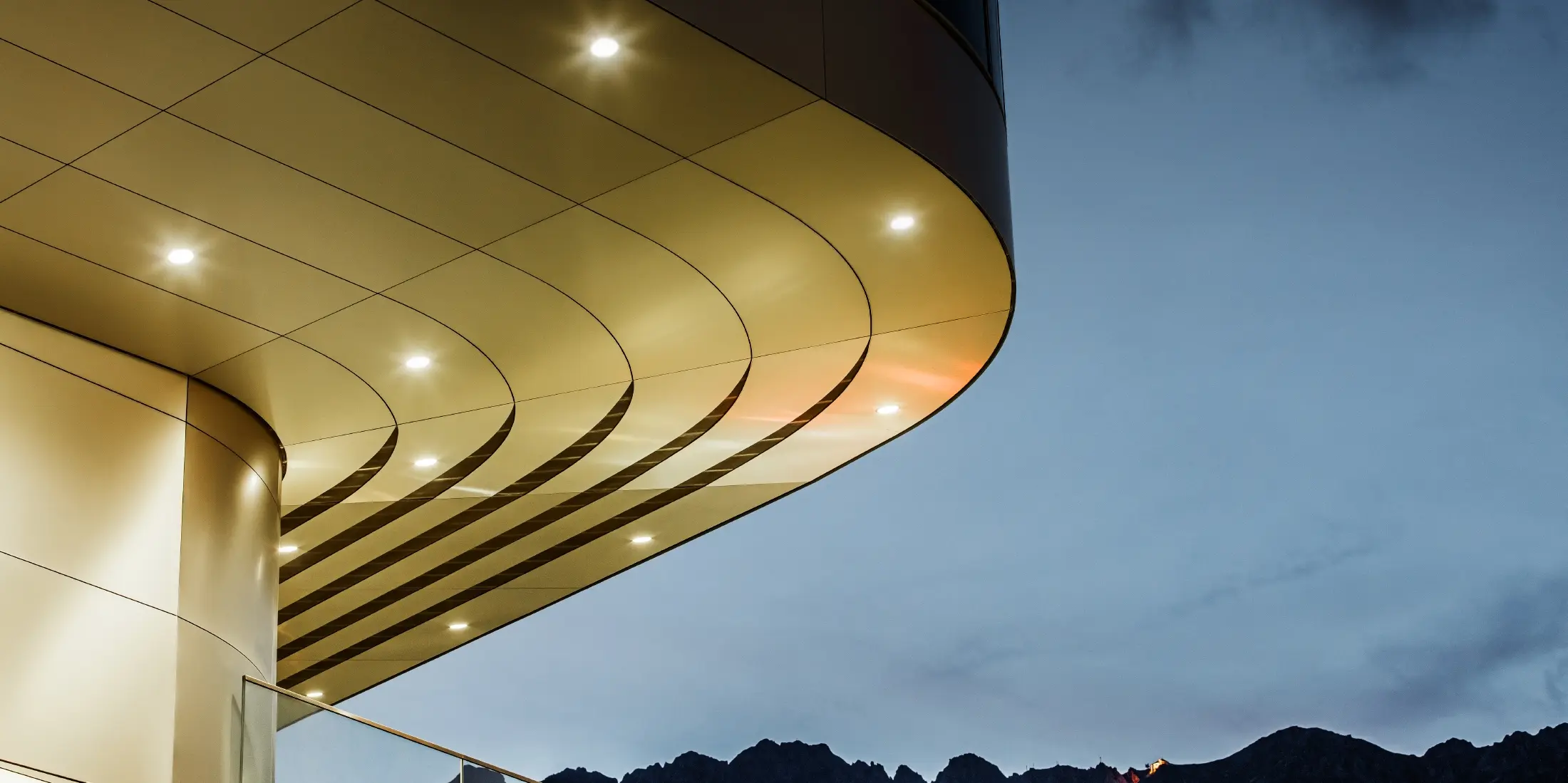 Billedet viser en imponerende arkitektonisk detalje af en PREFA PEMA-kompositplade af børstet aluminium. Pladen er let buet og har elegante, flydende linjer, der fremhæves særligt om aftenen ved hjælp af målrettet belysning. Pladens klare struktur og elegante aluminiumlook supplerer facadeens moderne design og tilbyder en elegant, moderne løsning til arkitektur af høj kvalitet.