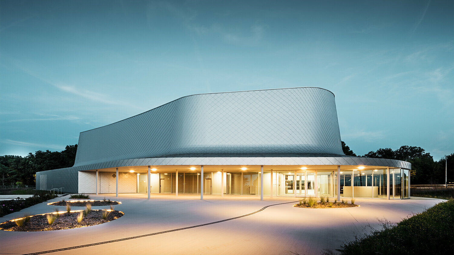 Seitliche Abendaufnahme der von Bohuon Bertic Architectes entworfenen Sporthalle in Basse-Goulaine mit Beleuchtung.