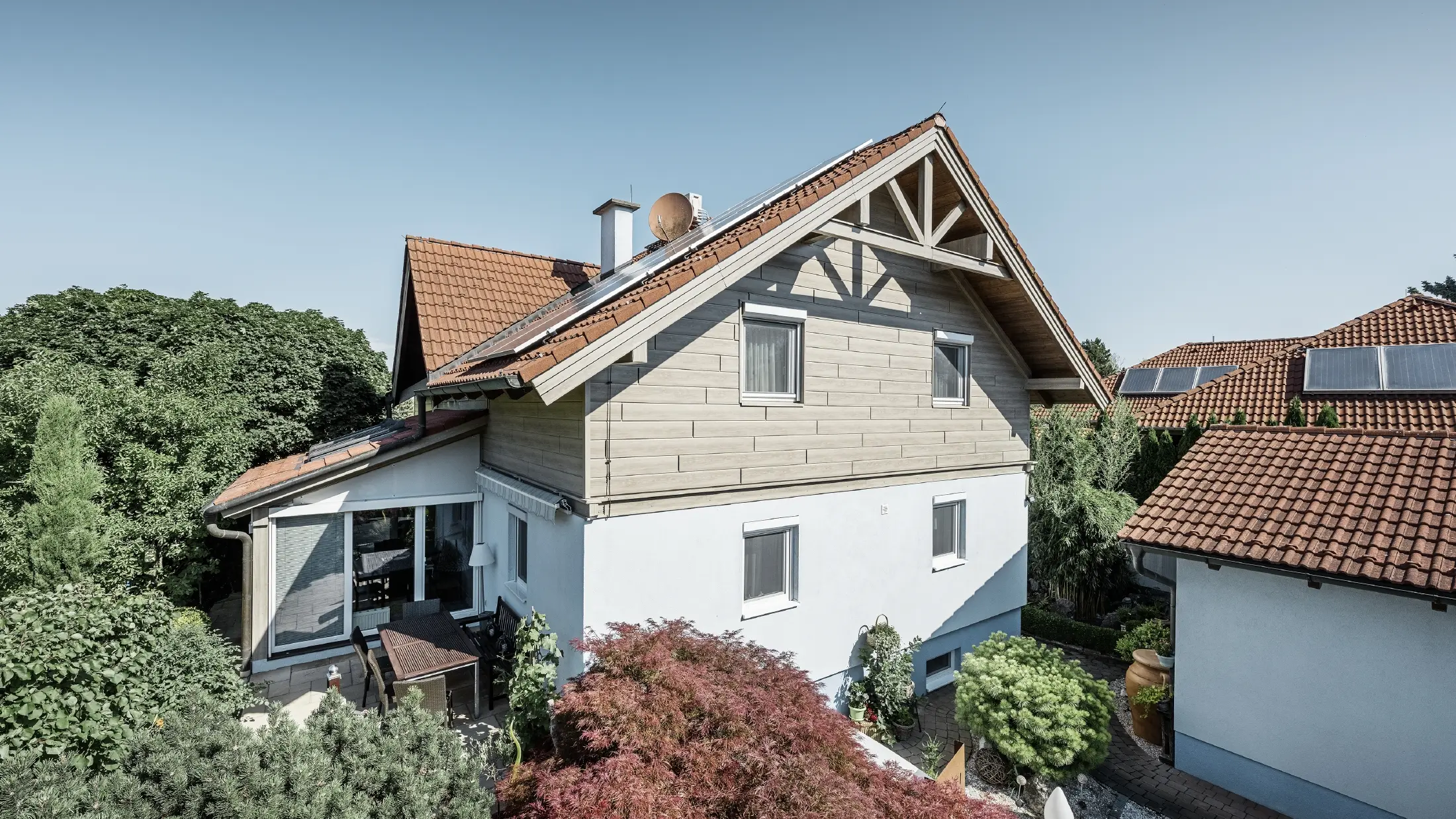 Enkeltfamiliehus med sadeltag og rødt tagdækning kombineret med et moderne facadedesign. Den øverste gavl er beklædt med grå eg PREFA-facadepaneler, mens stueetagen har en hvid pudset facade. Der er solcellemodul på taget. Huset ligger i en grøn have med terrasse, havemøbler og frodig beplantning, omgivet af andre boliger med lignende tagarkitektur.