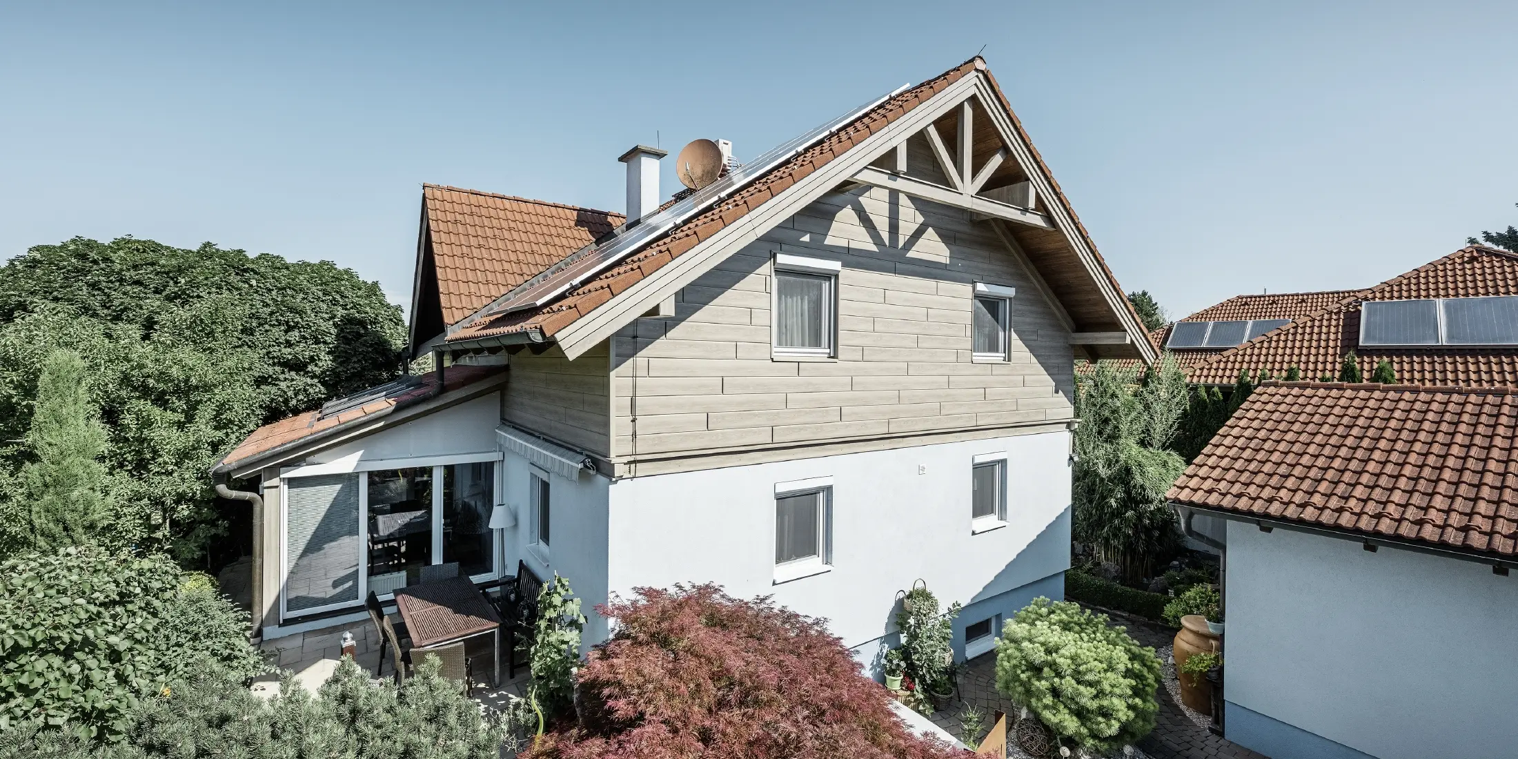 Enkeltfamiliehus med sadeltag og rødt tagdækning kombineret med et moderne facadedesign. Den øverste gavl er beklædt med grå eg PREFA-facadepaneler, mens stueetagen har en hvid pudset facade. Der er solcellemodul på taget. Huset ligger i en grøn have med terrasse, havemøbler og frodig beplantning, omgivet af andre boliger med lignende tagarkitektur.